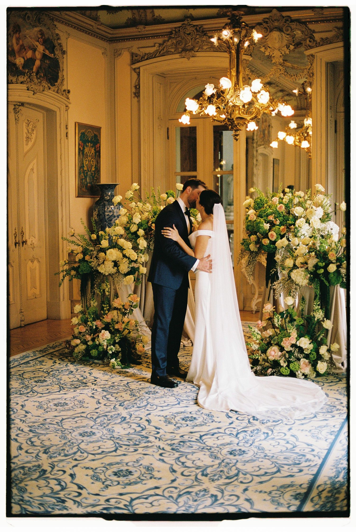 Bride and groom share first kiss surrounded by lush floral arrangements in an elegant chandelier-lit ballroom.