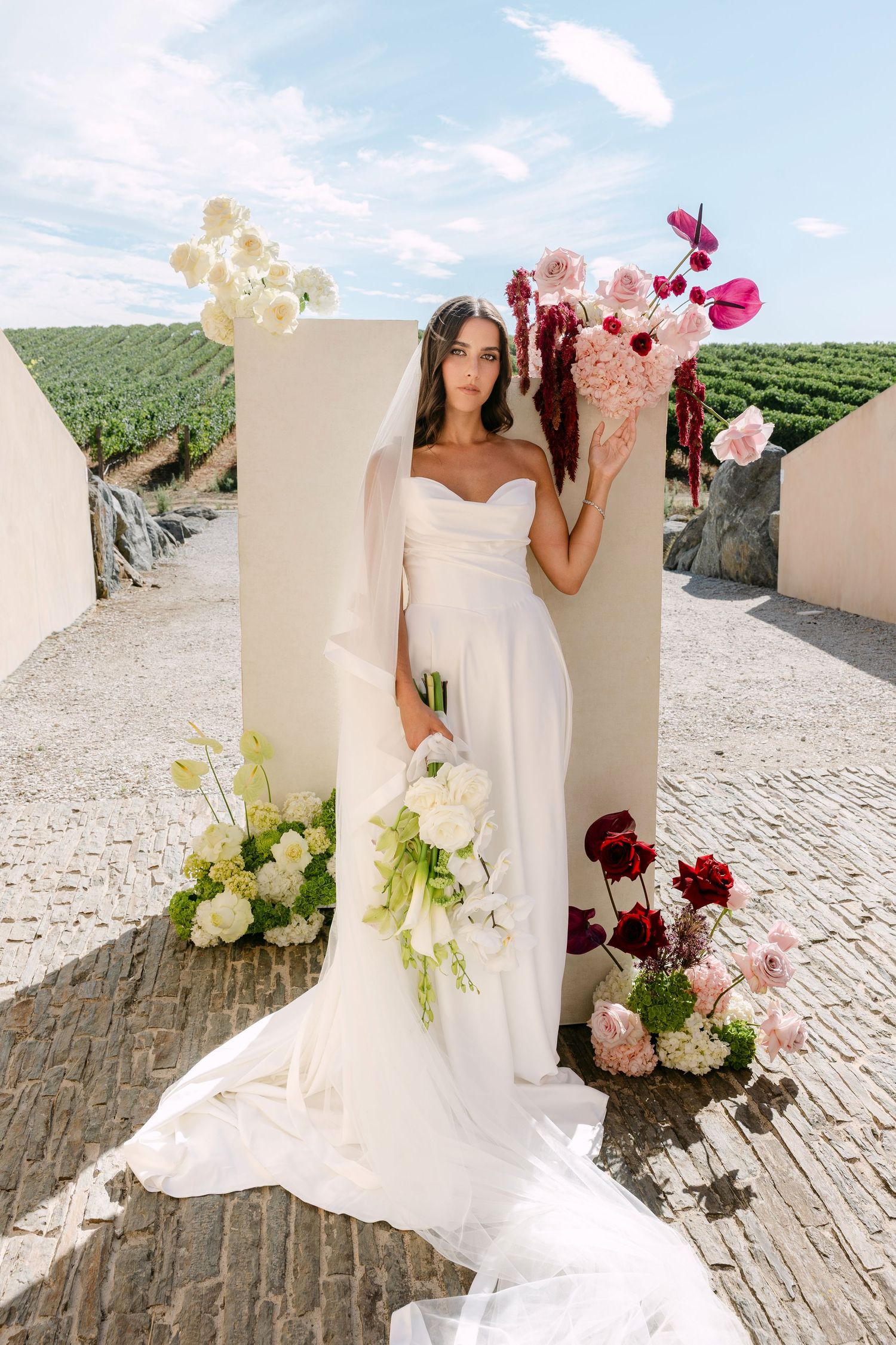 Bride in white off-shoulder gown holding floral bouquet stands before white panels with vineyard backdrop.