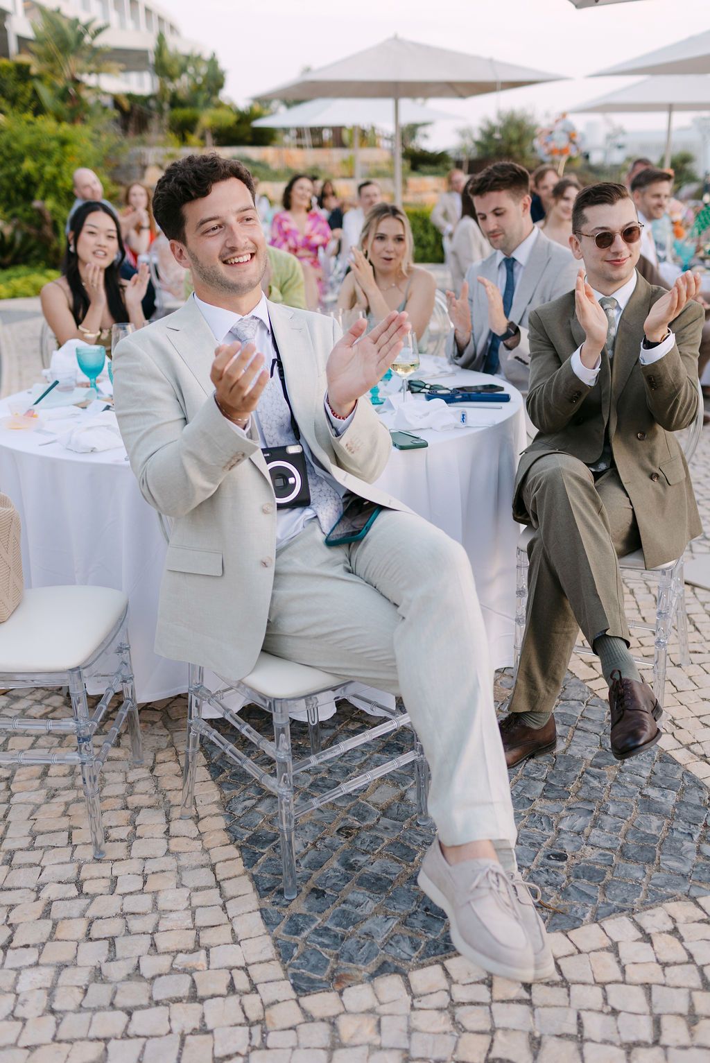 Wedding guests in formal attire seated and clapping during outdoor celebration.