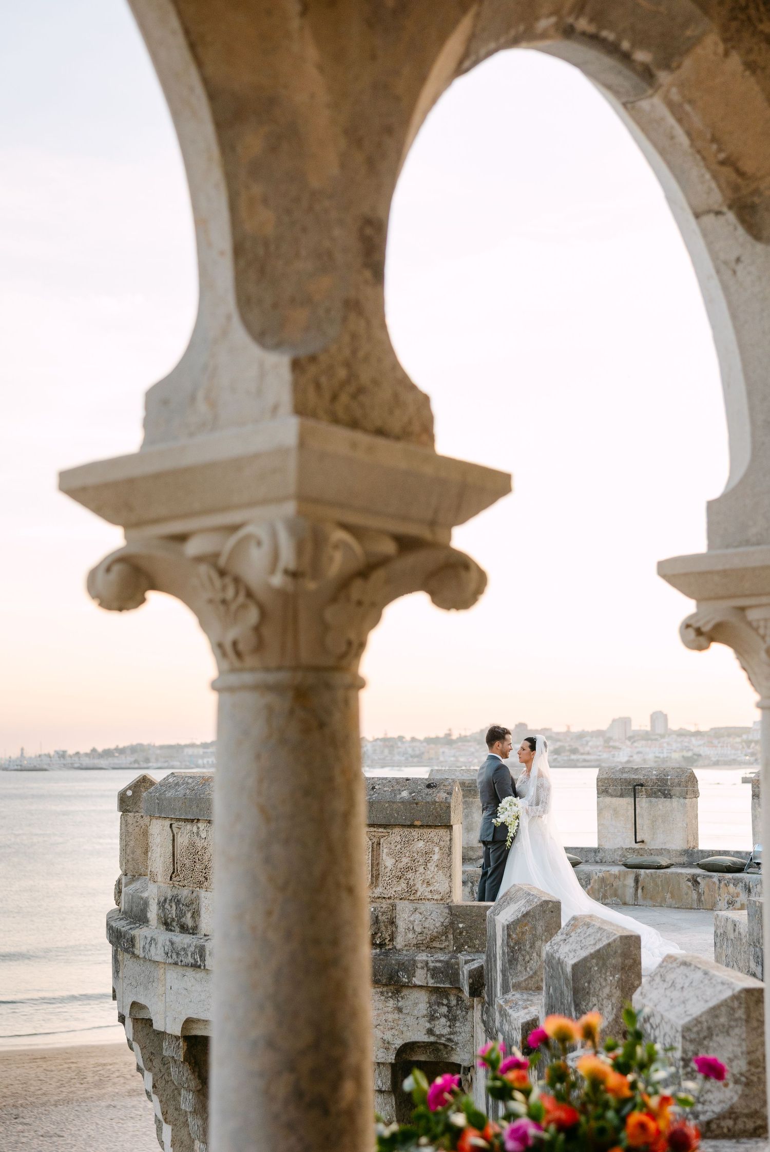 Romantic wedding portrait framed by stone archways overlooking waterfront at golden hour.