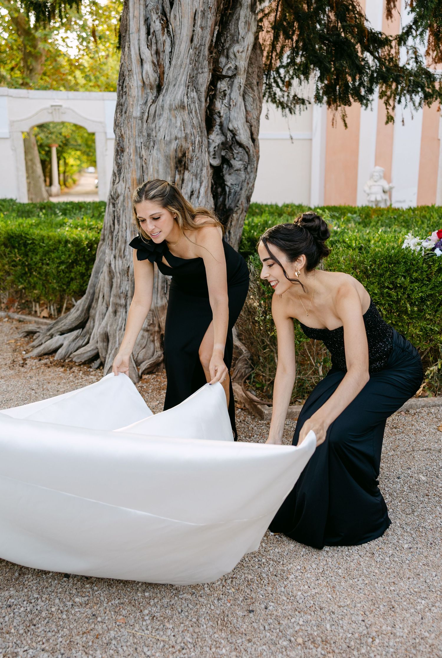 Bridesmaids in black dresses help arrange the bride's wedding gown train outdoors.