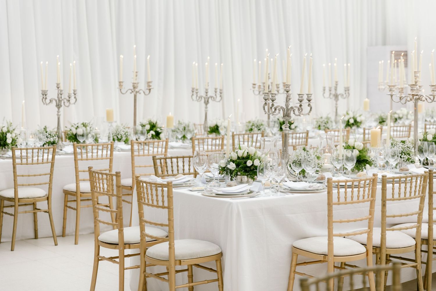 Elegant wedding reception setup with white linens, gold chiavari chairs, and crystal chandeliers.