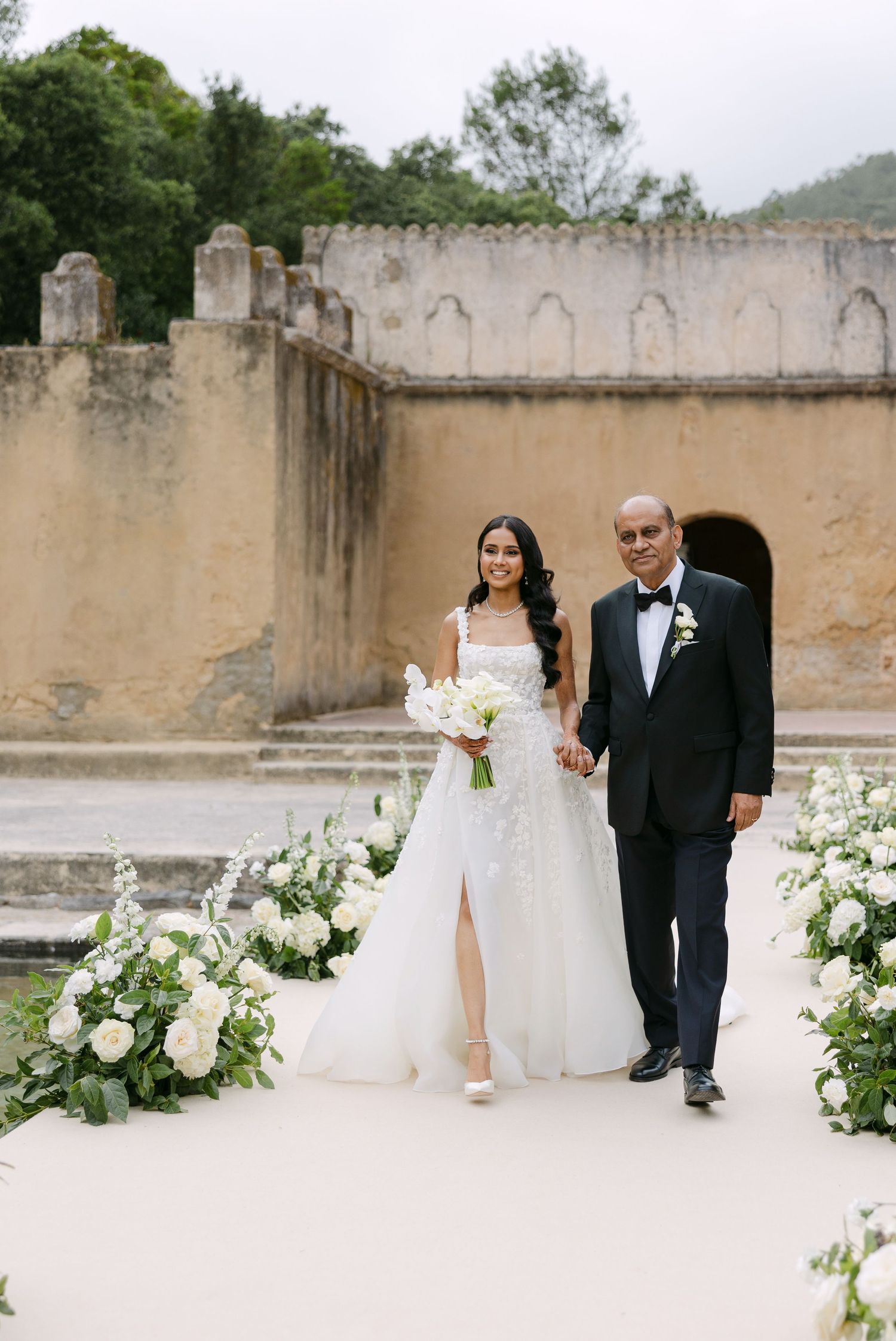 A couple walks along a flower-lined path at their wedding venue featuring historic stone architecture.