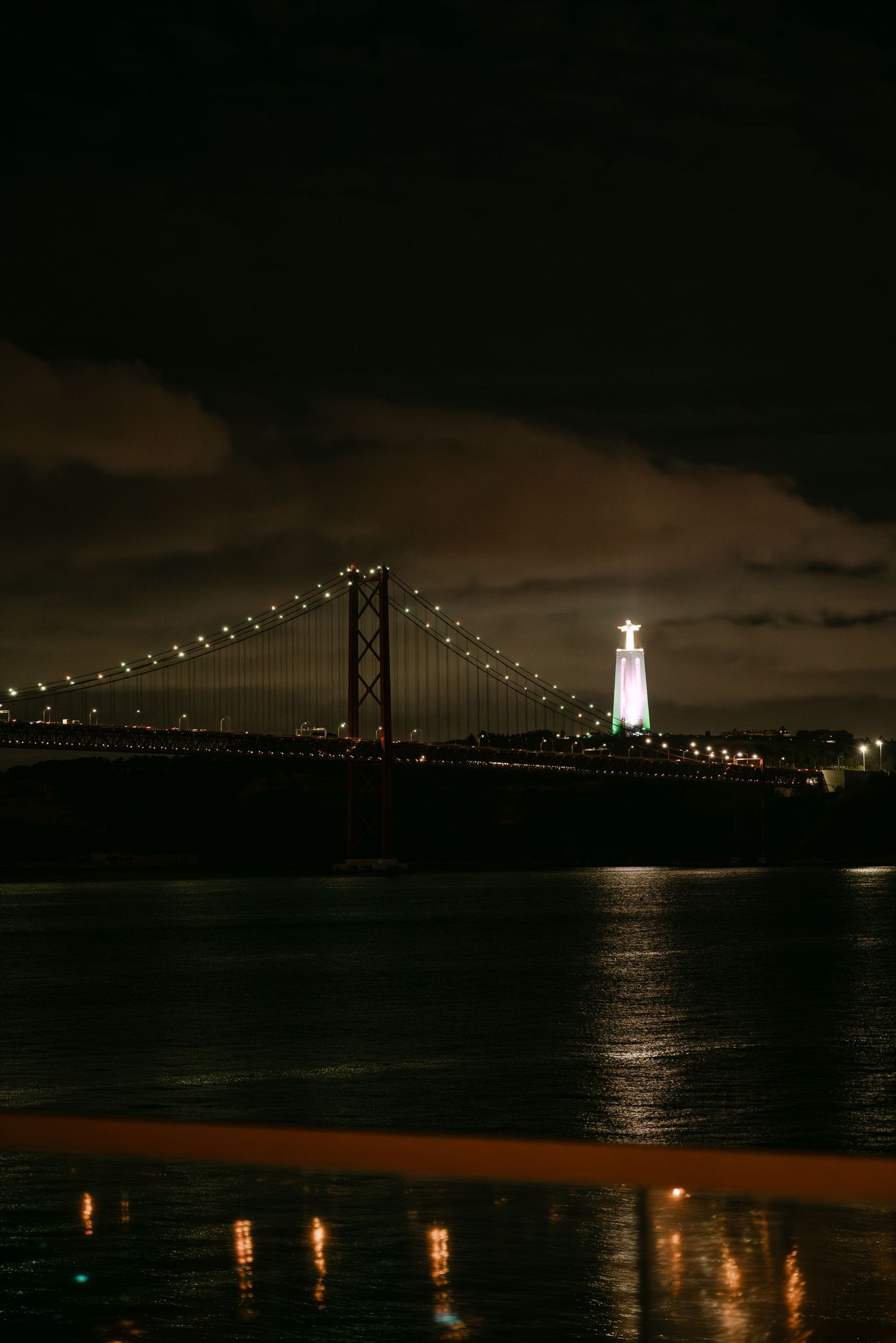 Night view of a bridge and lighthouse with illuminated lights reflecting on calm water.