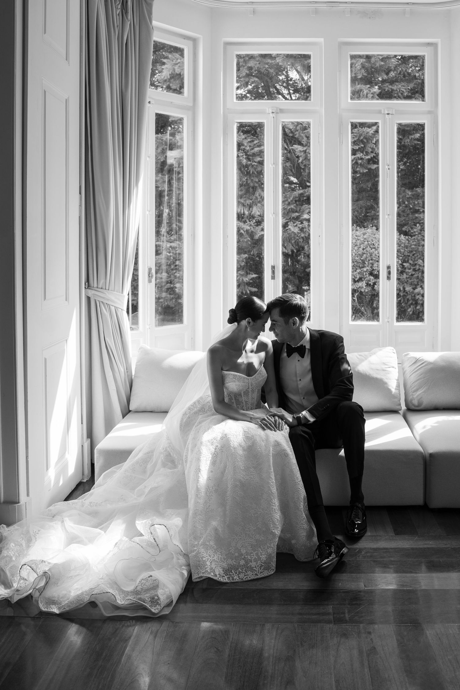 Black and white wedding photo series showing intimate moments in a sunlit room with large windows.