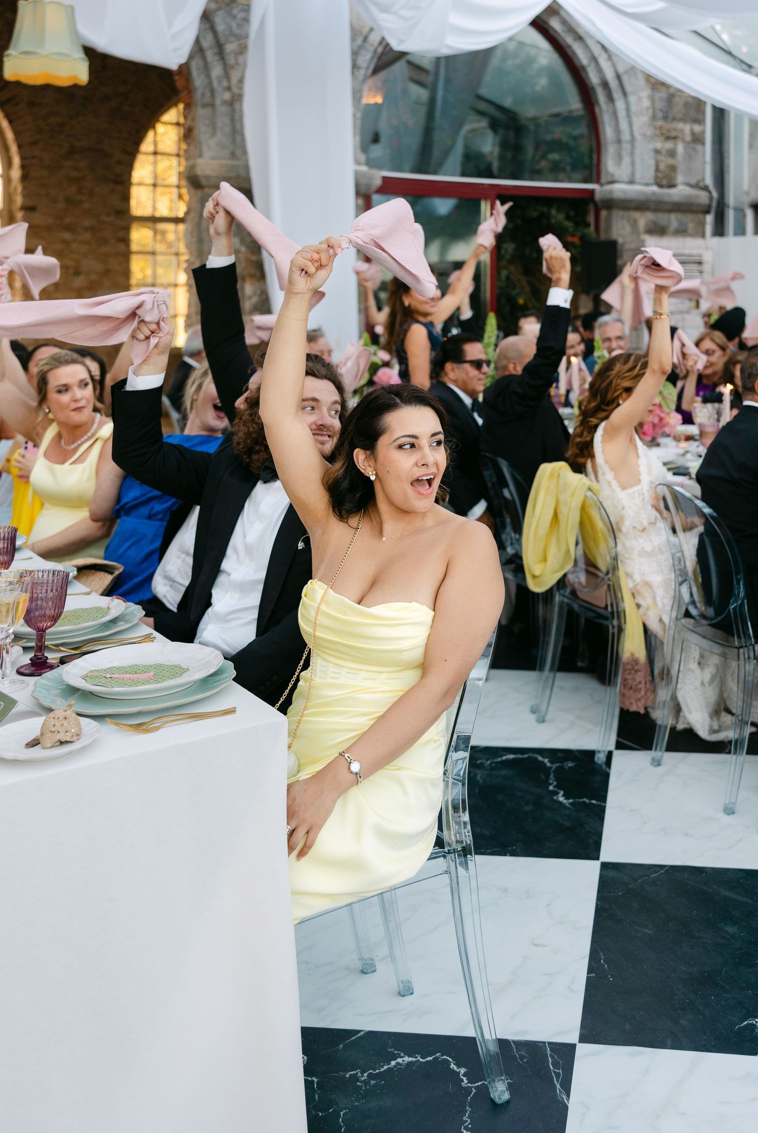 Joyful wedding guests in formal attire celebrate with raised arms at a black and white checkered venue.