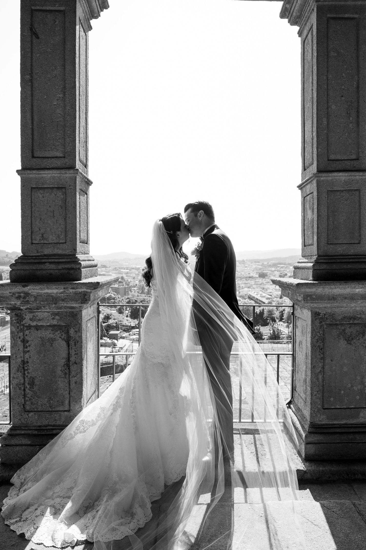 Elegant bride and groom share intimate moment on balcony with columned architecture.