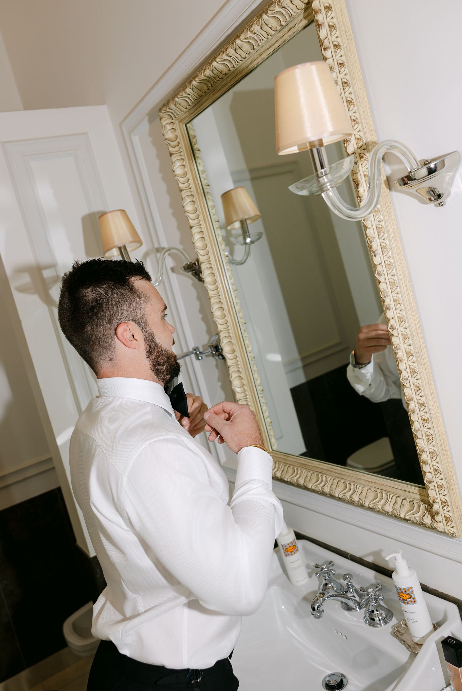 Getting ready scene in bathroom with ornate gold-framed mirror and white fixtures.