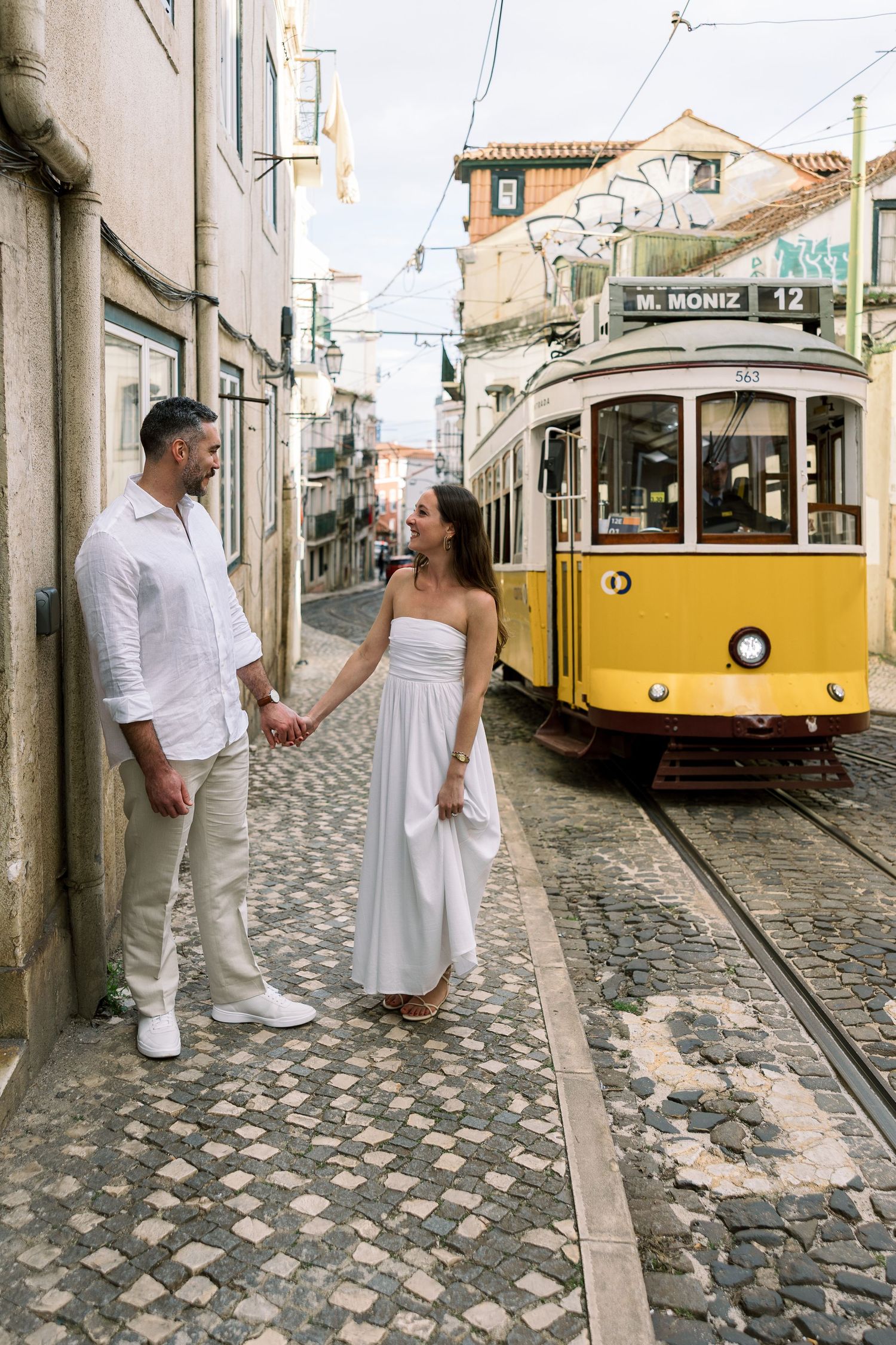 Couple in white outfits holding hands on cobblestone street with iconic yellow tram 12 in Lisbon, Portugal.