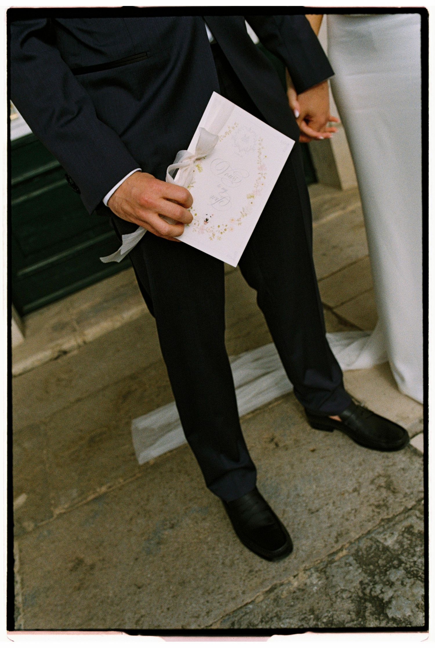 Groom in black suit holds white wedding program with ribbon outside church steps.