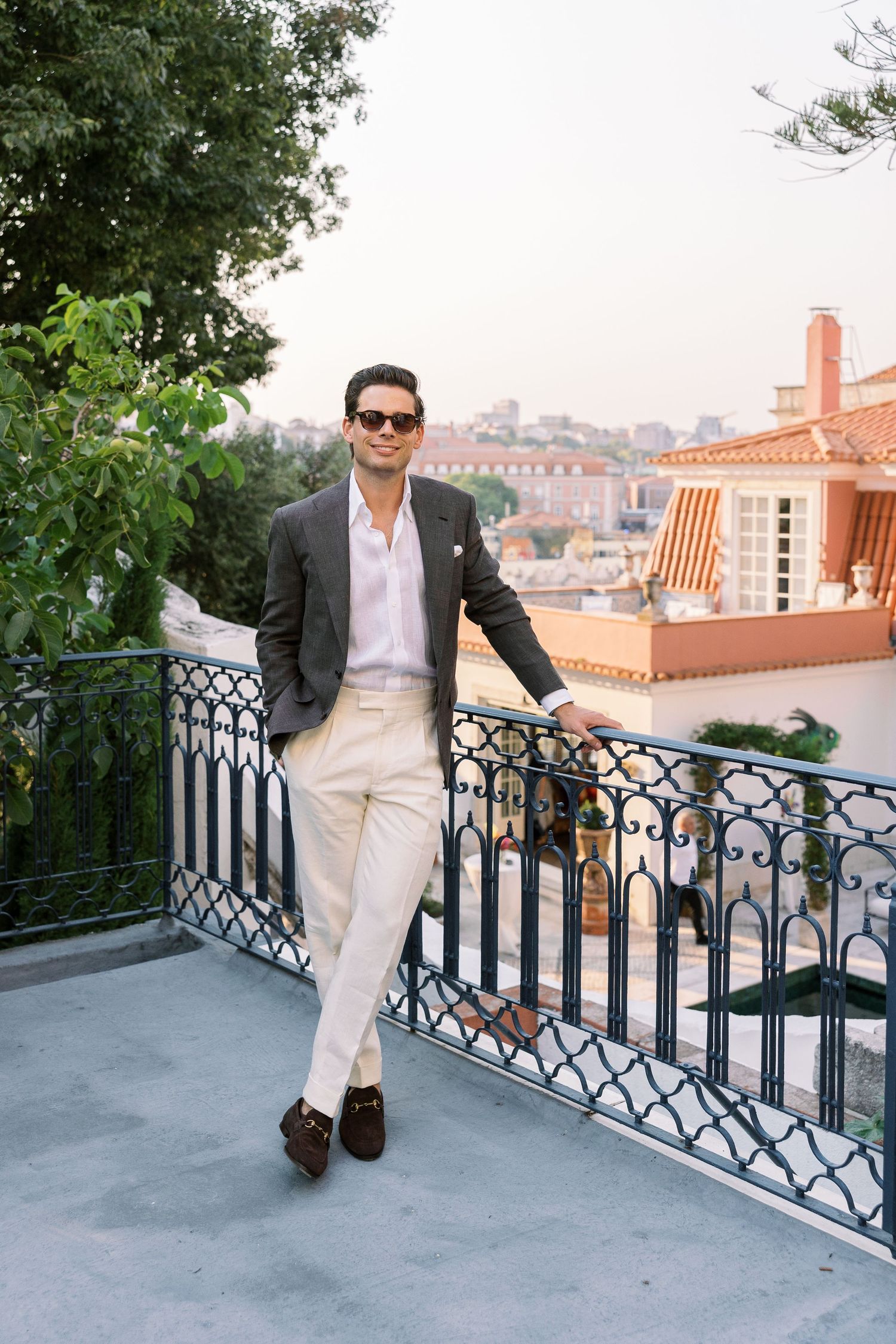 Stylish man in blazer and cream trousers leaning on iron balcony railing with city rooftop views.