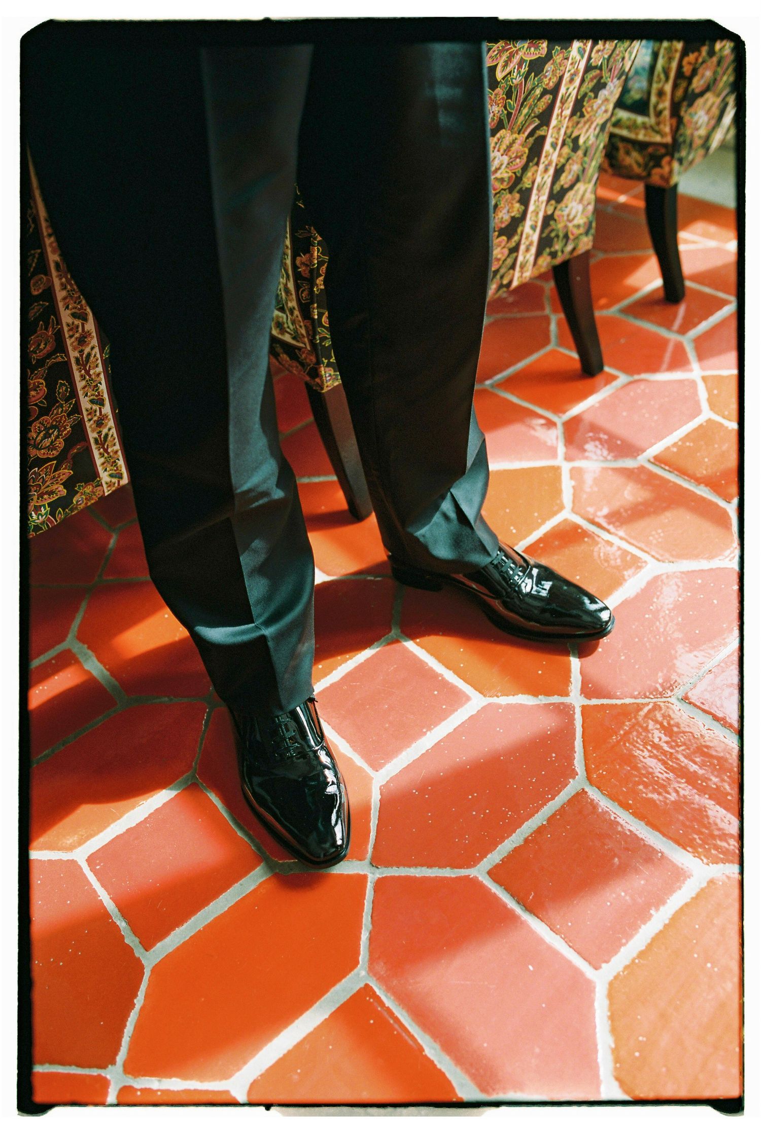 Man in black tuxedo pants and patent leather dress shoes standing on red hexagonal tile floor.