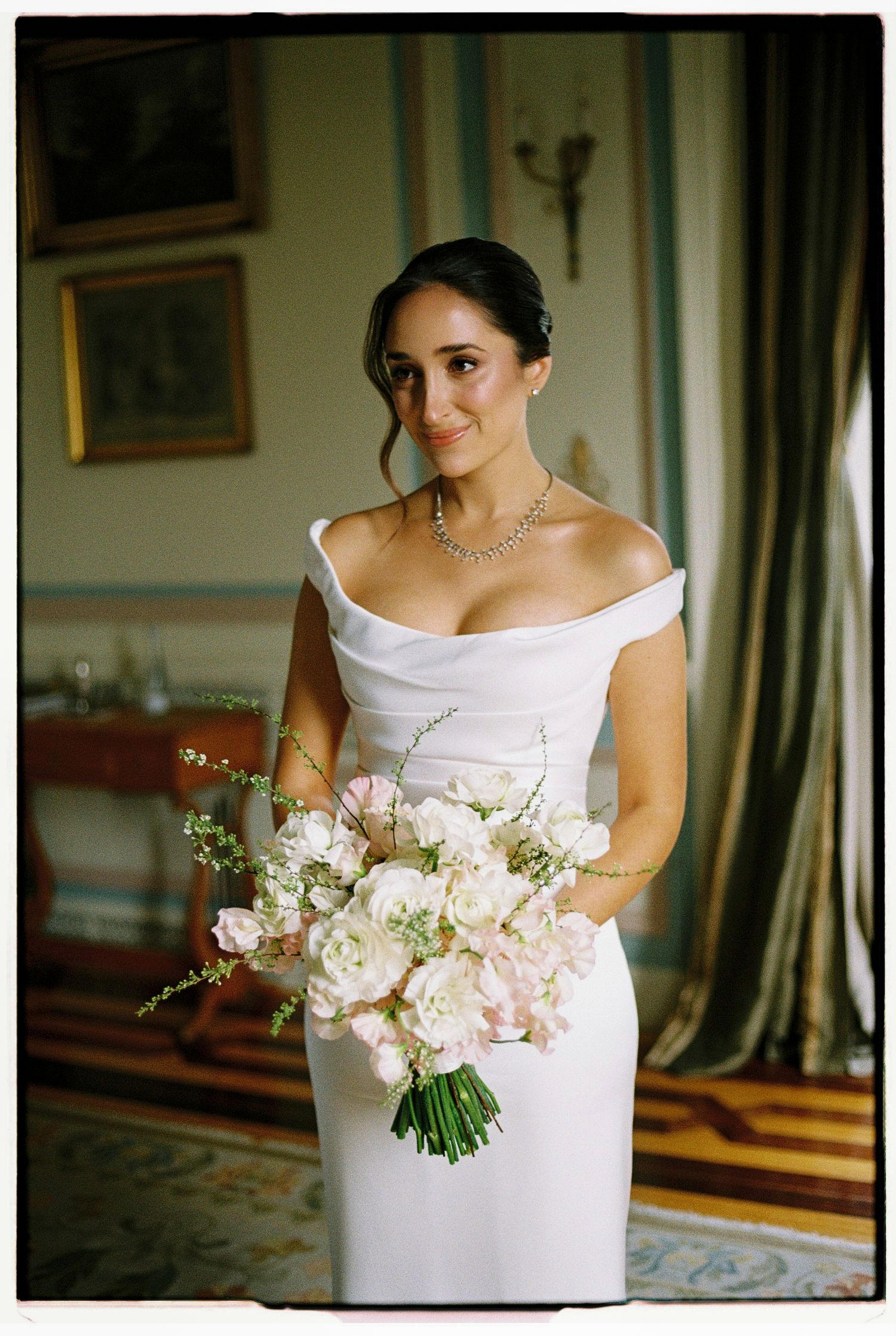 Bride in off-shoulder white gown holding lush pink and white floral bouquet, wearing diamond necklace in elegant room.
