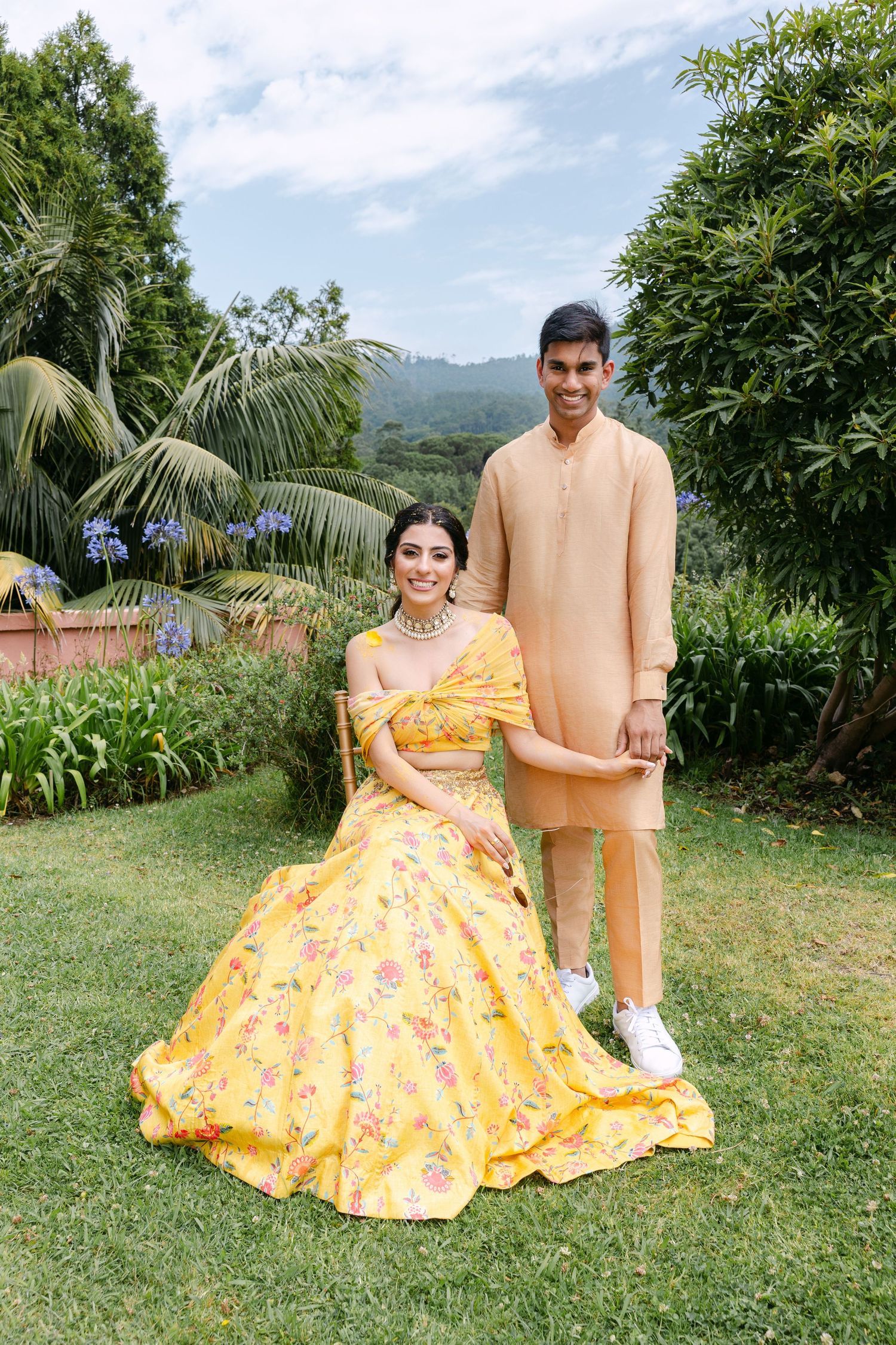 A couple in traditional Indian wedding attire poses in a garden, wearing yellow and peach outfits.