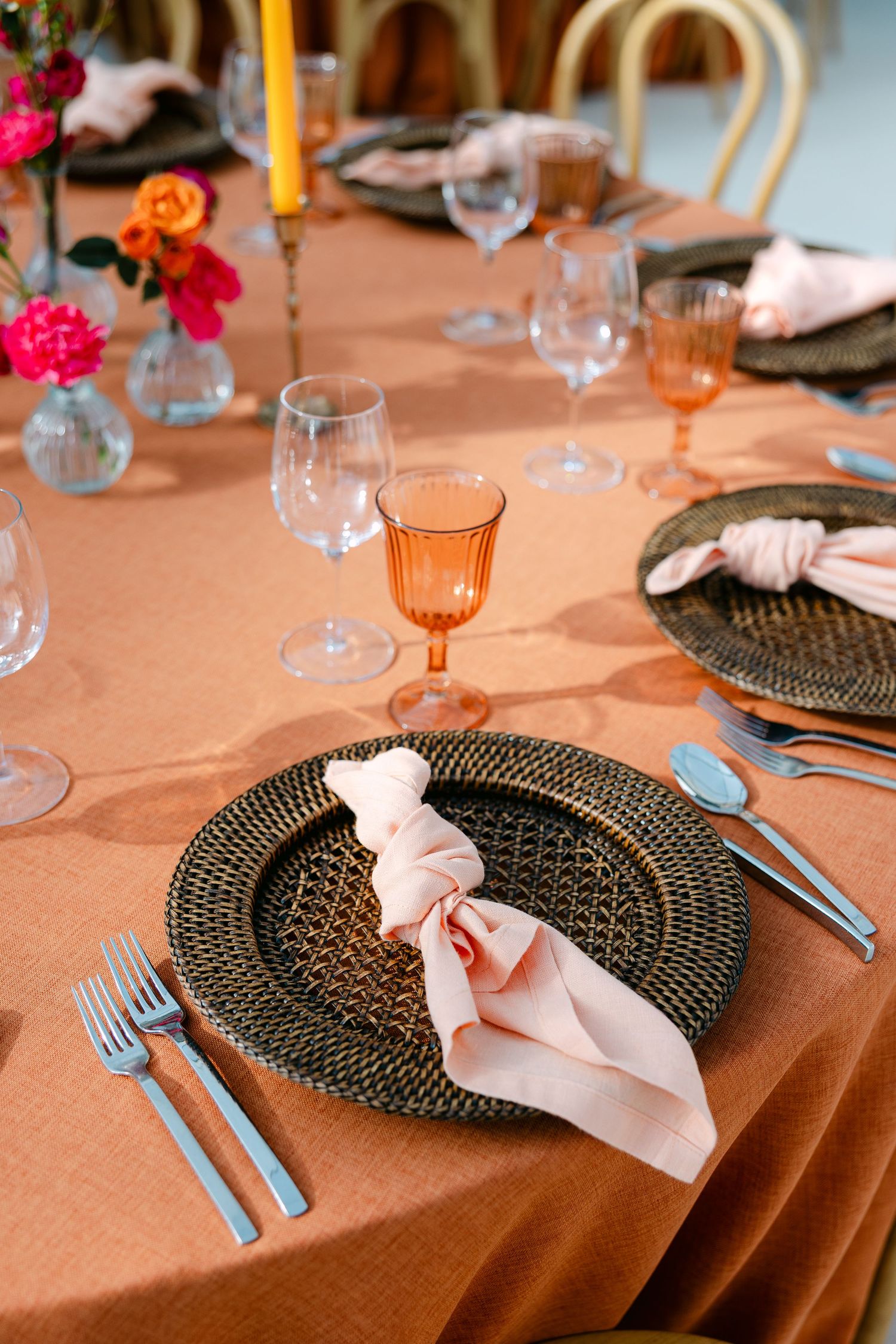 Elegant table settings with dark woven placemats, pink napkins, and amber glassware on a peach-colored tablecloth.