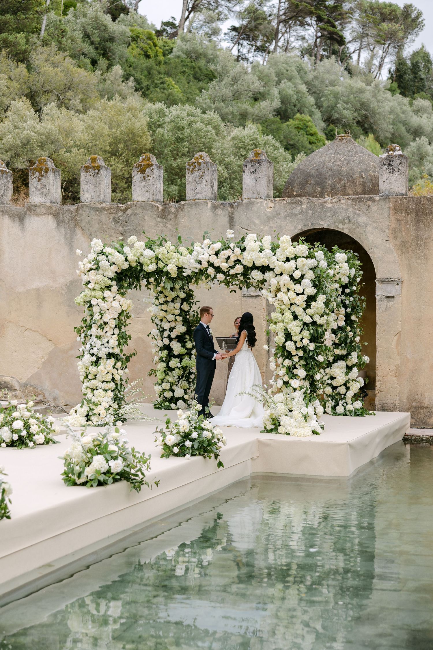 A wedding ceremony setup with white floral arch and reflecting pool against ancient stone walls.