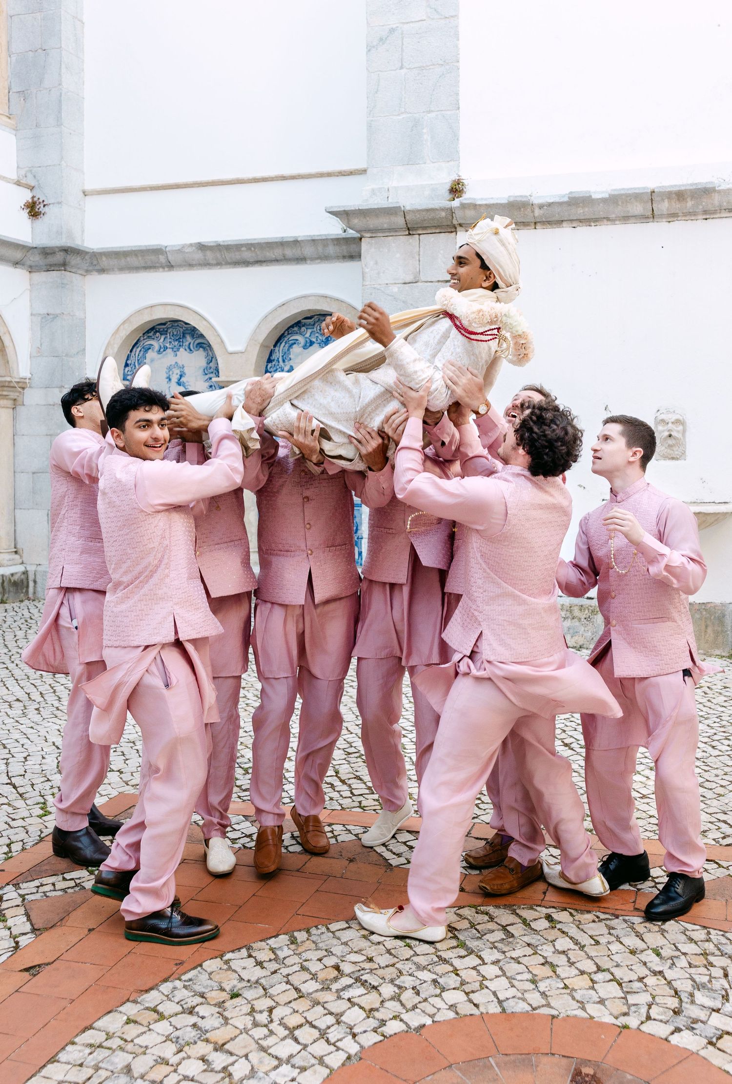 Groomsmen in matching pink suits posing playfully against white building wall.