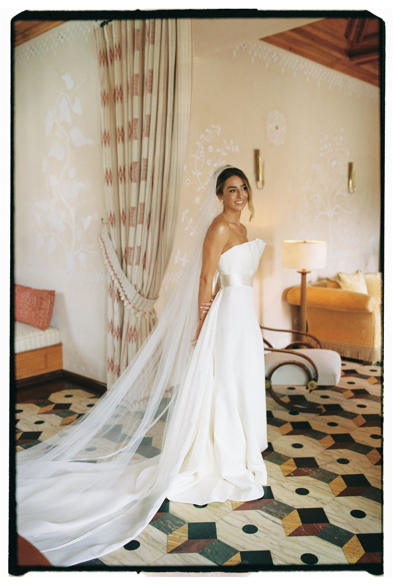 Bride in strapless white gown with long veil stands elegantly in a decorated hotel room with patterned floor.