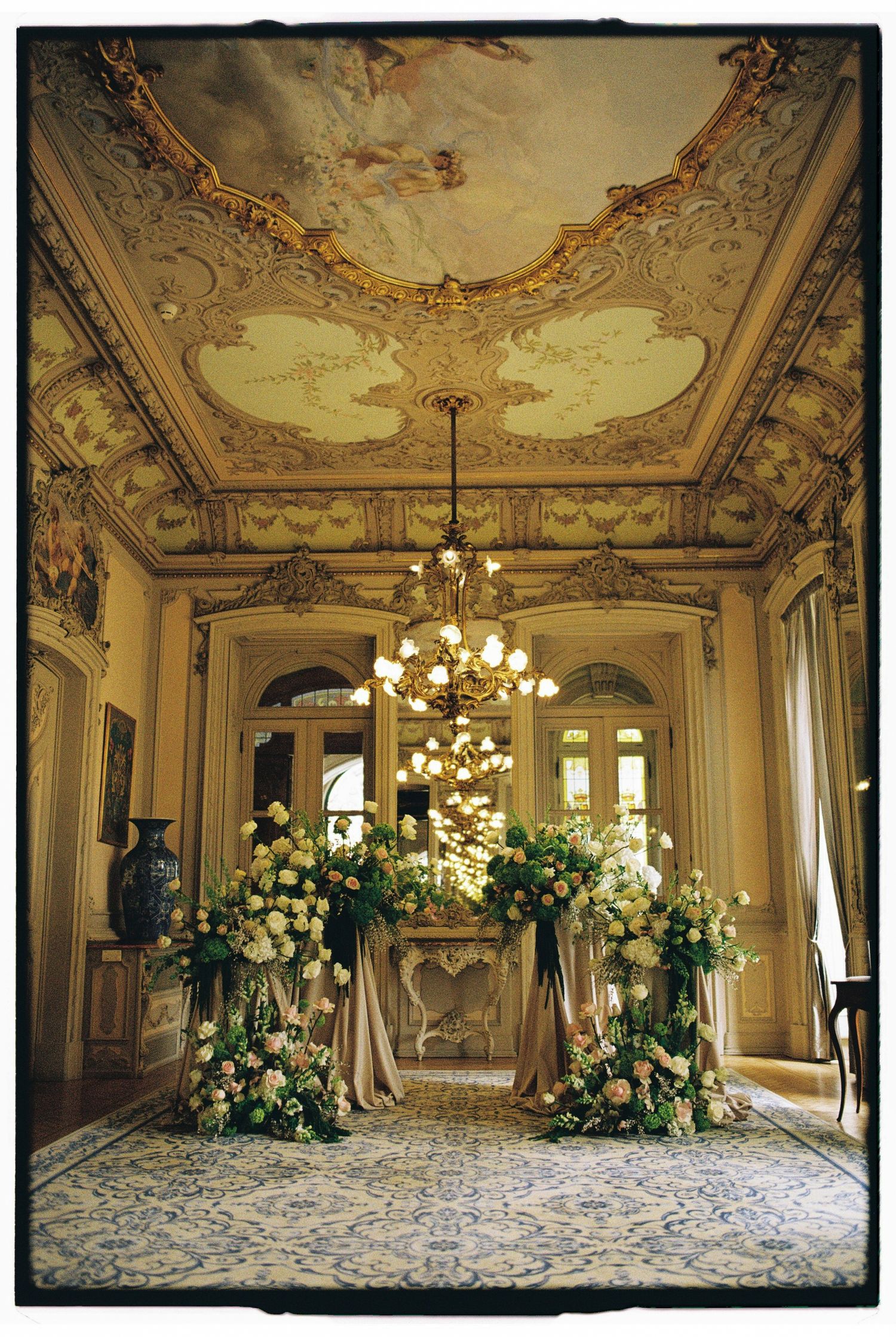 Ornate baroque ballroom with gilded ceiling, chandelier, and lush floral arch arrangement for an elegant wedding ceremony.