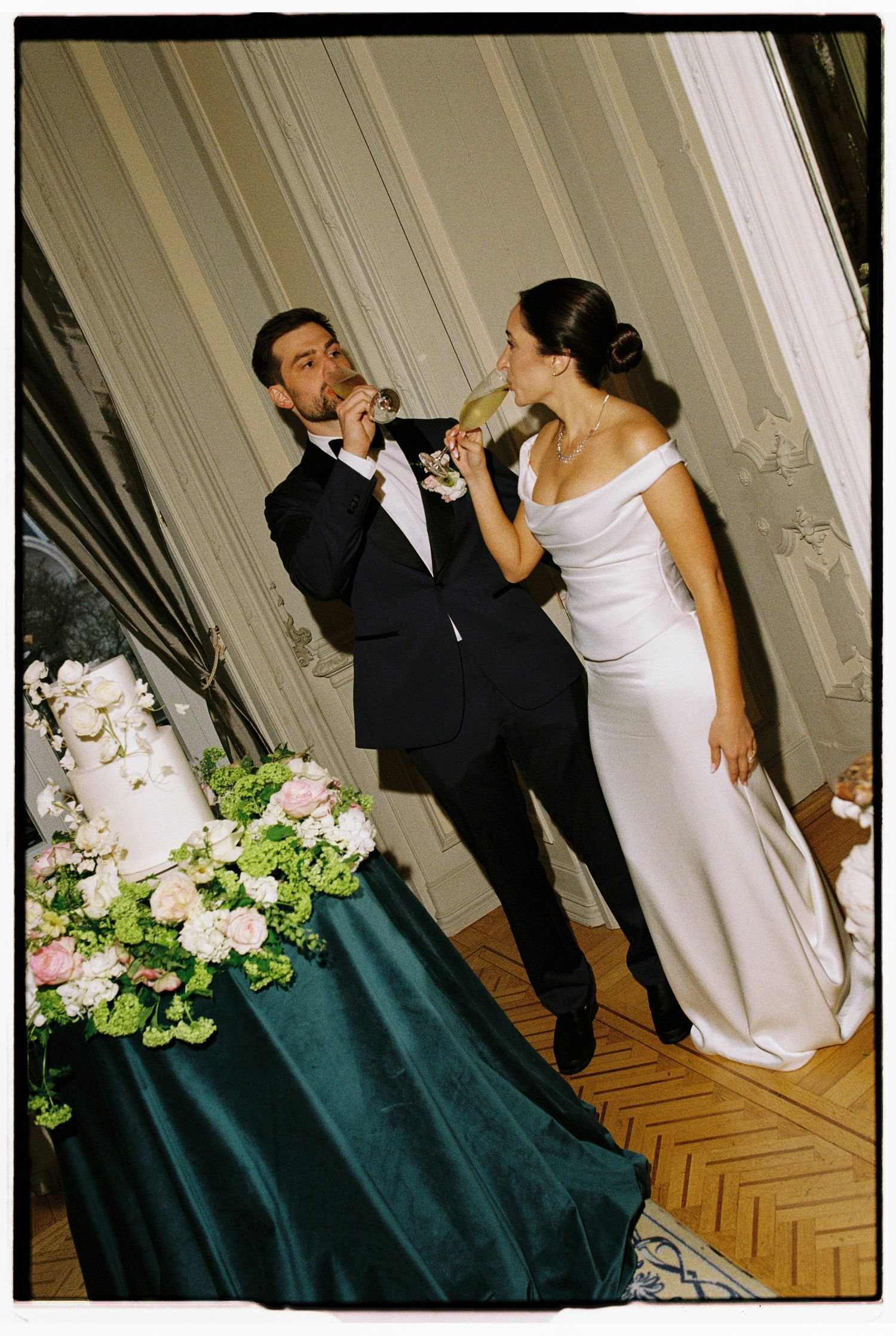 Bride and groom toasting with champagne beside a floral wedding cake at an elegant reception venue.
