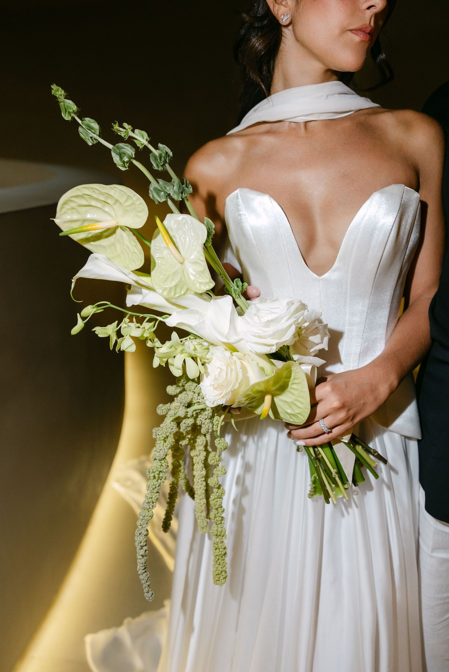Bride in deep V strapless white gown holds elegant white anthurium and rose bouquet with trailing greenery.