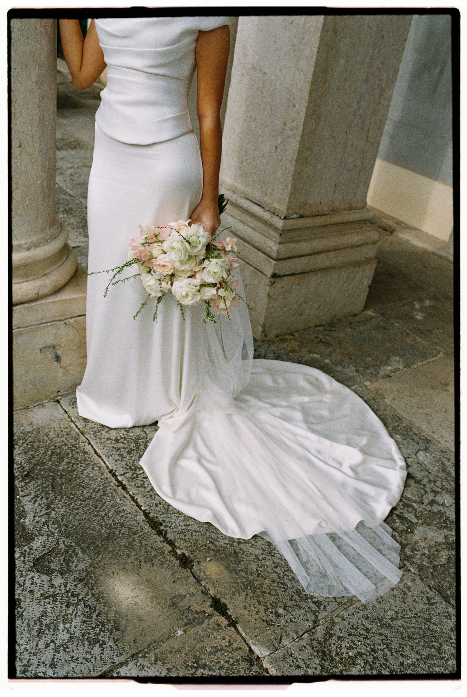 Bride in sleek white gown holds a soft pink and white bouquet beside stone columns, veil trailing.