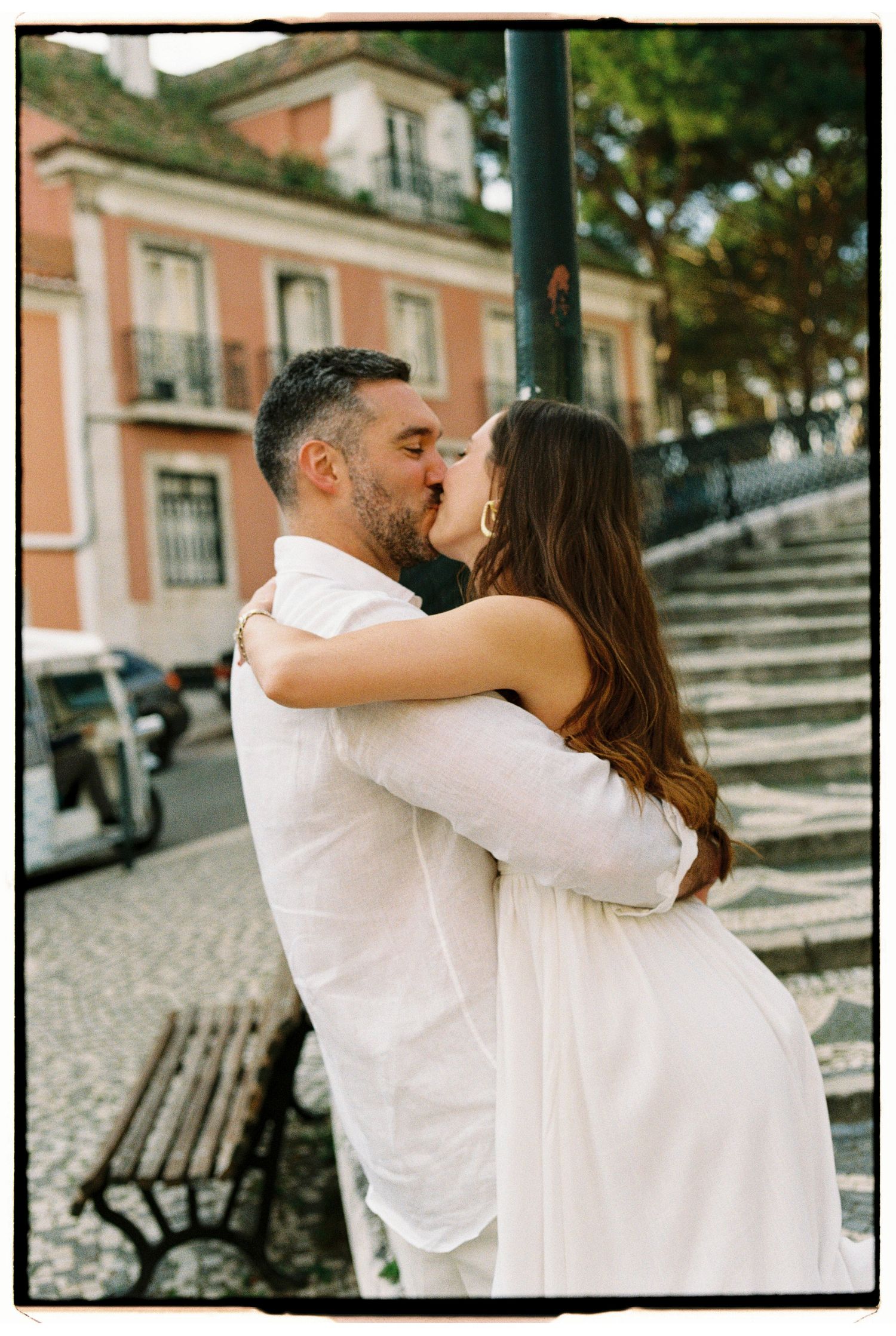 Couple sharing a romantic kiss outdoors in Lisbon, dressed in white, with cobblestone streets and pink building behind.