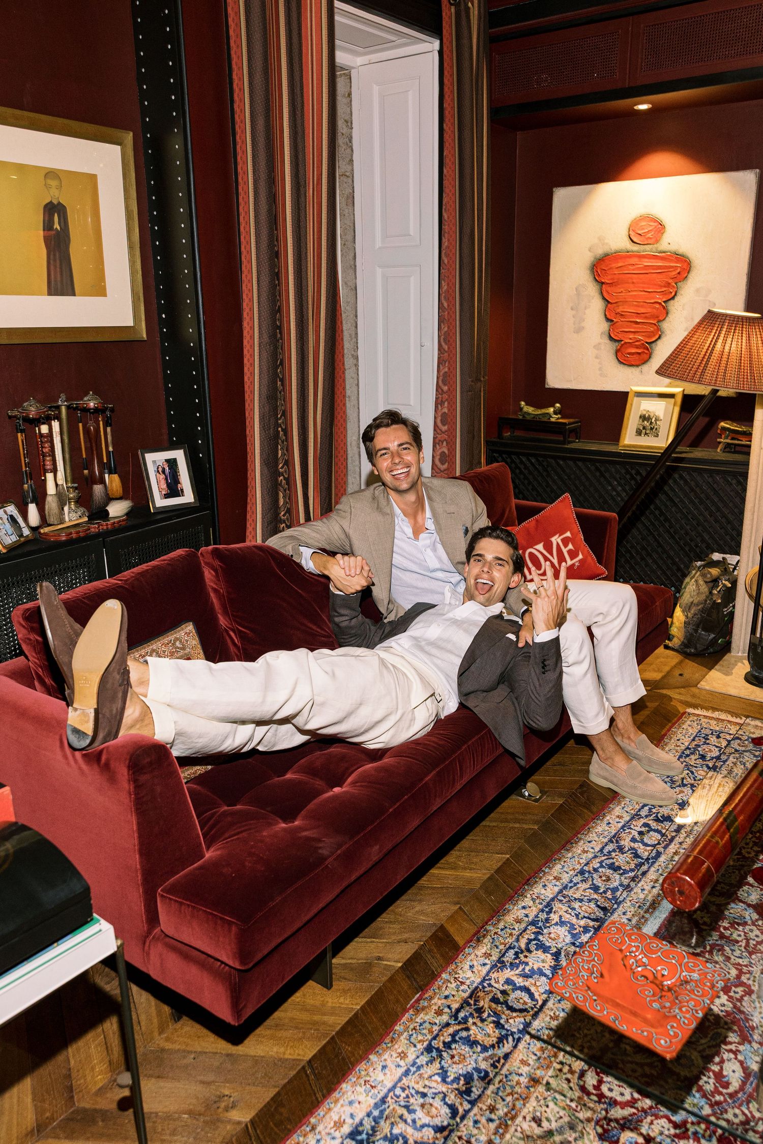 Two men lounging on a red velvet tufted sofa in an elegantly decorated room with artwork and warm lighting.