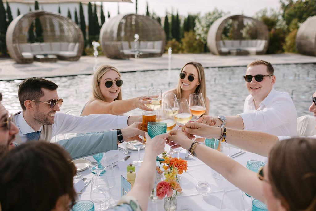 Wedding guests raise glasses in a celebratory toast at an outdoor reception table.
