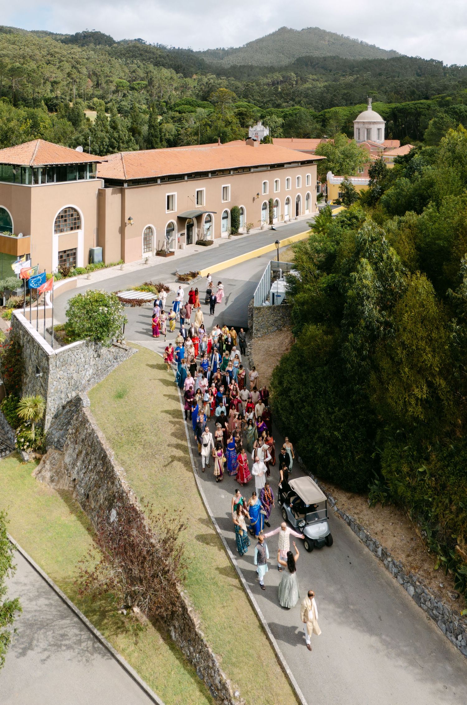 Aerial view of wedding guests gathered along a stone wall path near buildings.