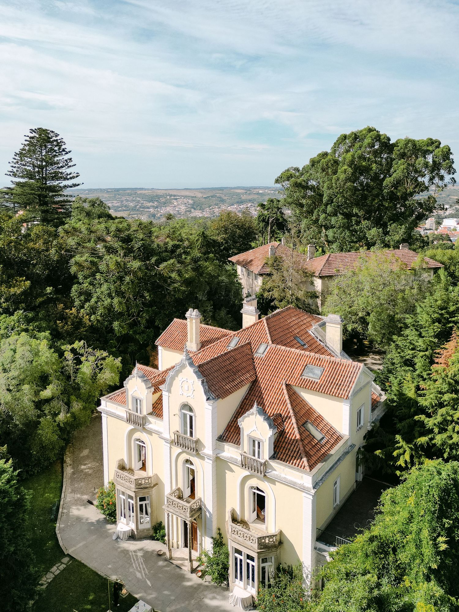 Aerial view of elegant cream-colored villa with terracotta roof tiles surrounded by lush greenery.