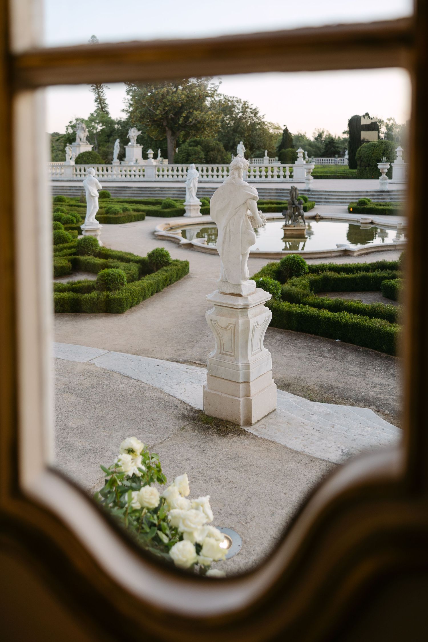 Elegant white garden statues and pathways viewed through ornate window frame.