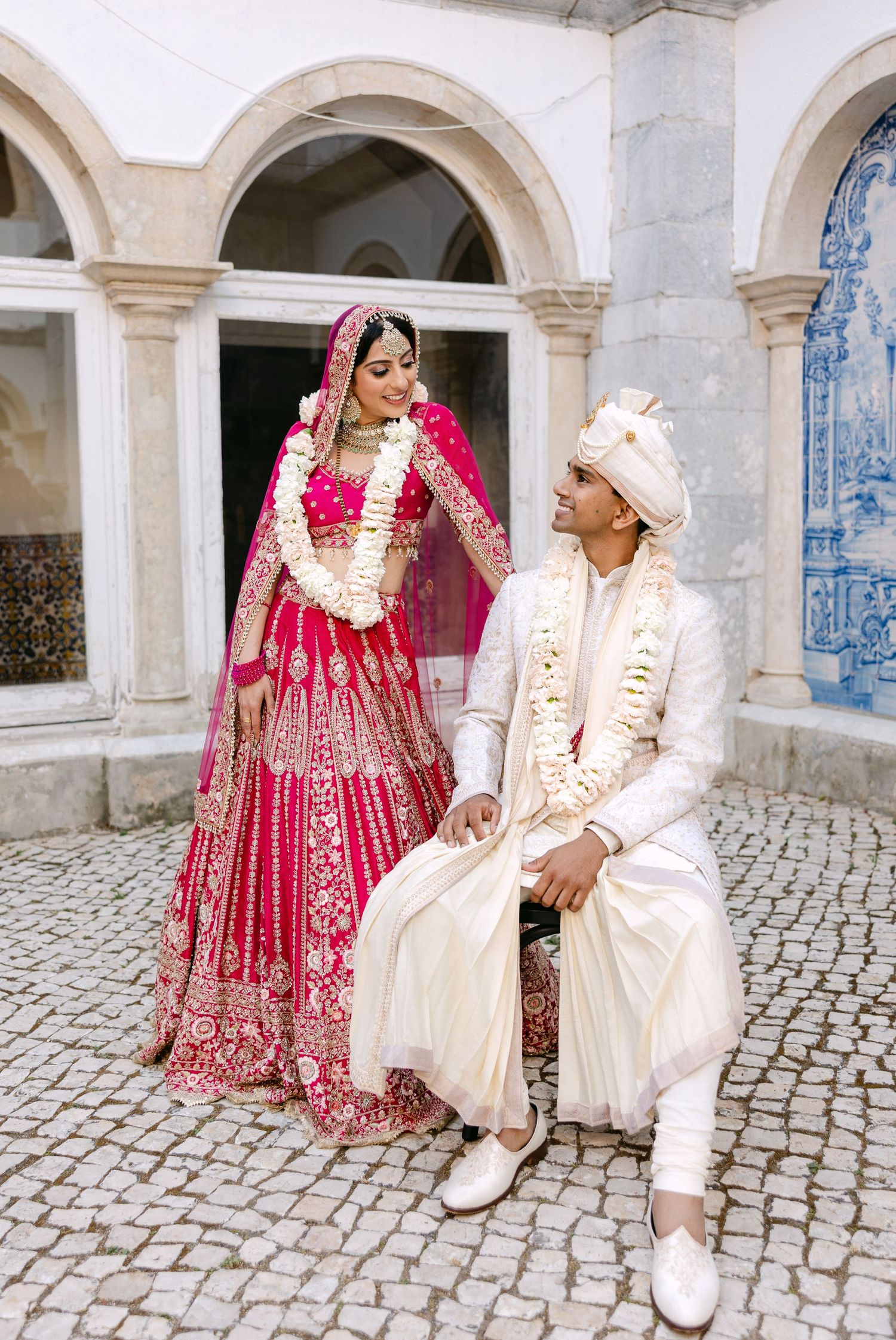 A couple in traditional Indian wedding attire poses in front of an arched colonnade.