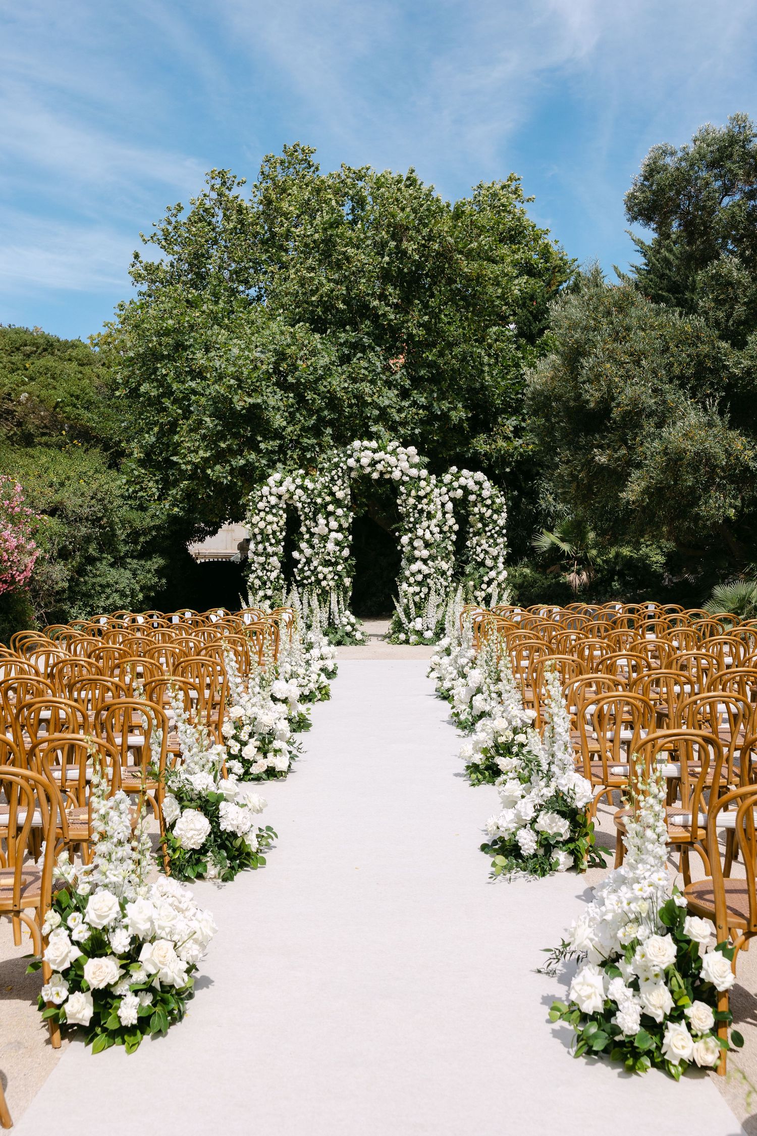 Outdoor wedding ceremony setup with white floral arch and wooden chairs under blue sky.