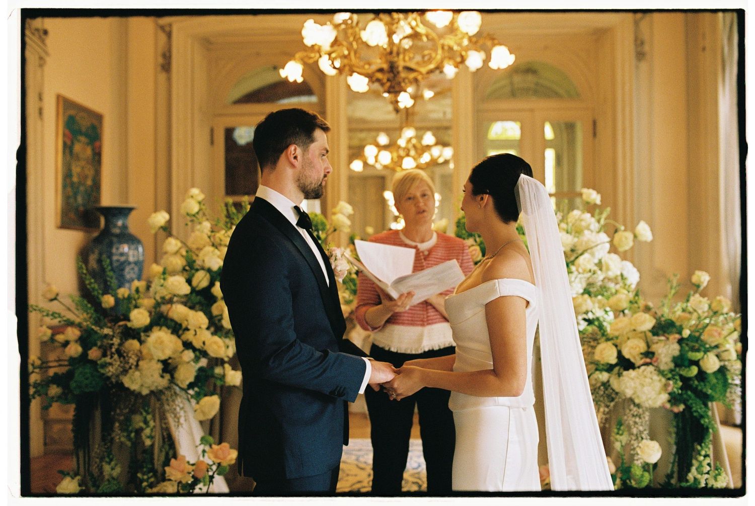 Elegant wedding ceremony indoors with couple exchanging vows before officiant, surrounded by lush floral arrangements and chandelier.