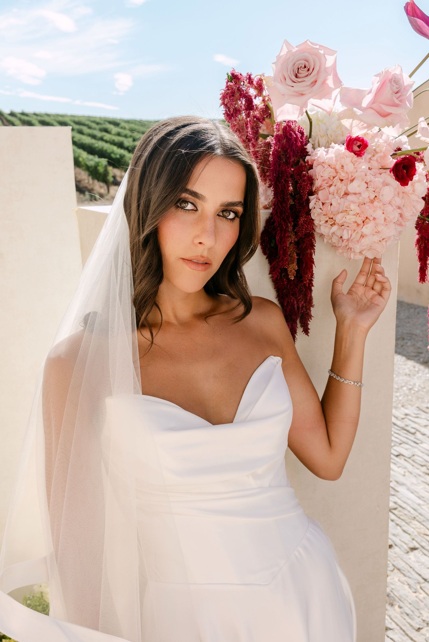 Elegant bride in strapless white gown and veil poses beside pink and red floral arrangement outdoors.