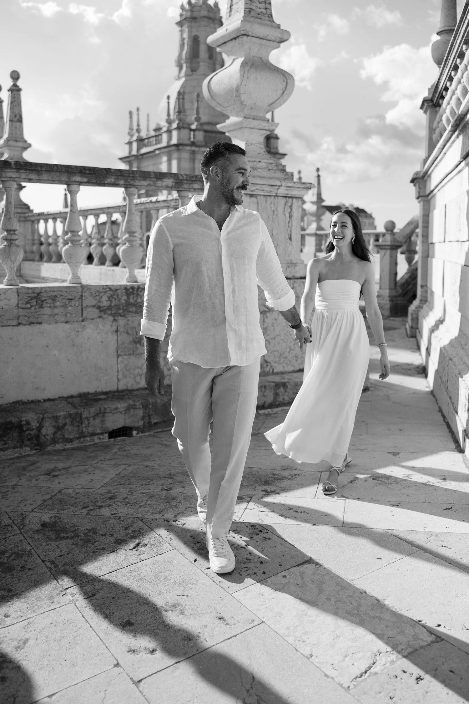 Black and white photo of a couple in white outfits holding hands while walking on a terrace of a historic building.