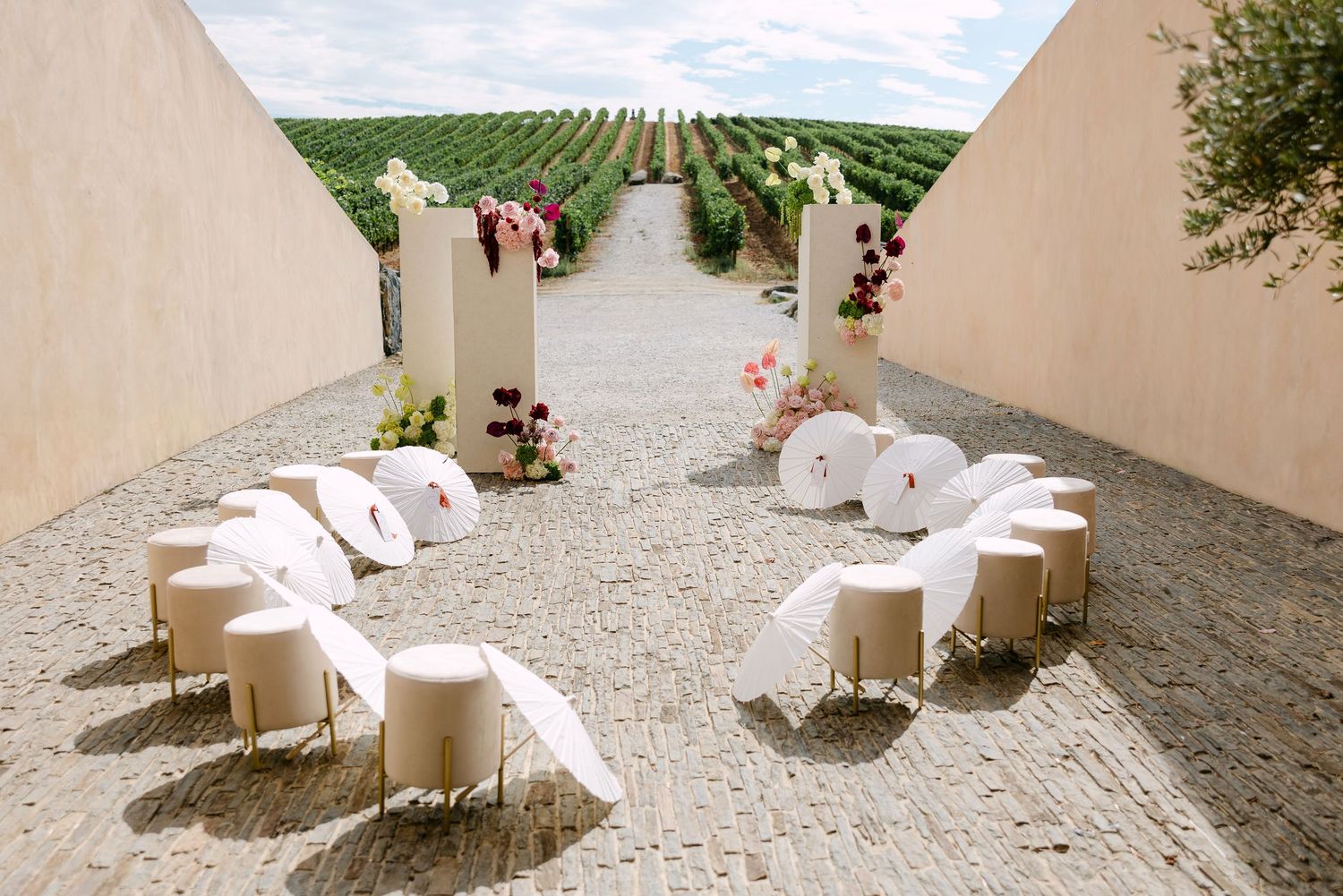 Elegant outdoor vineyard wedding ceremony setup with white parasols, cylindrical seats, and floral arch decor.