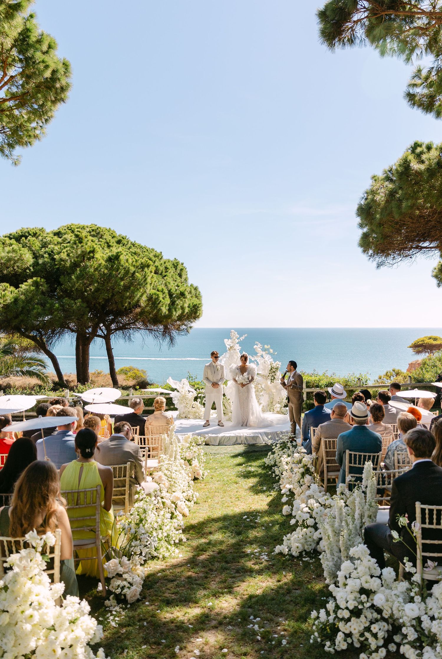 Outdoor wedding ceremony setup with white floral arrangements and chairs overlooking the ocean on a sunny day.