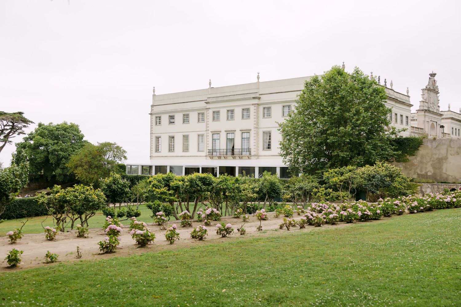 A grand white mansion sits atop a manicured lawn with garden landscaping.