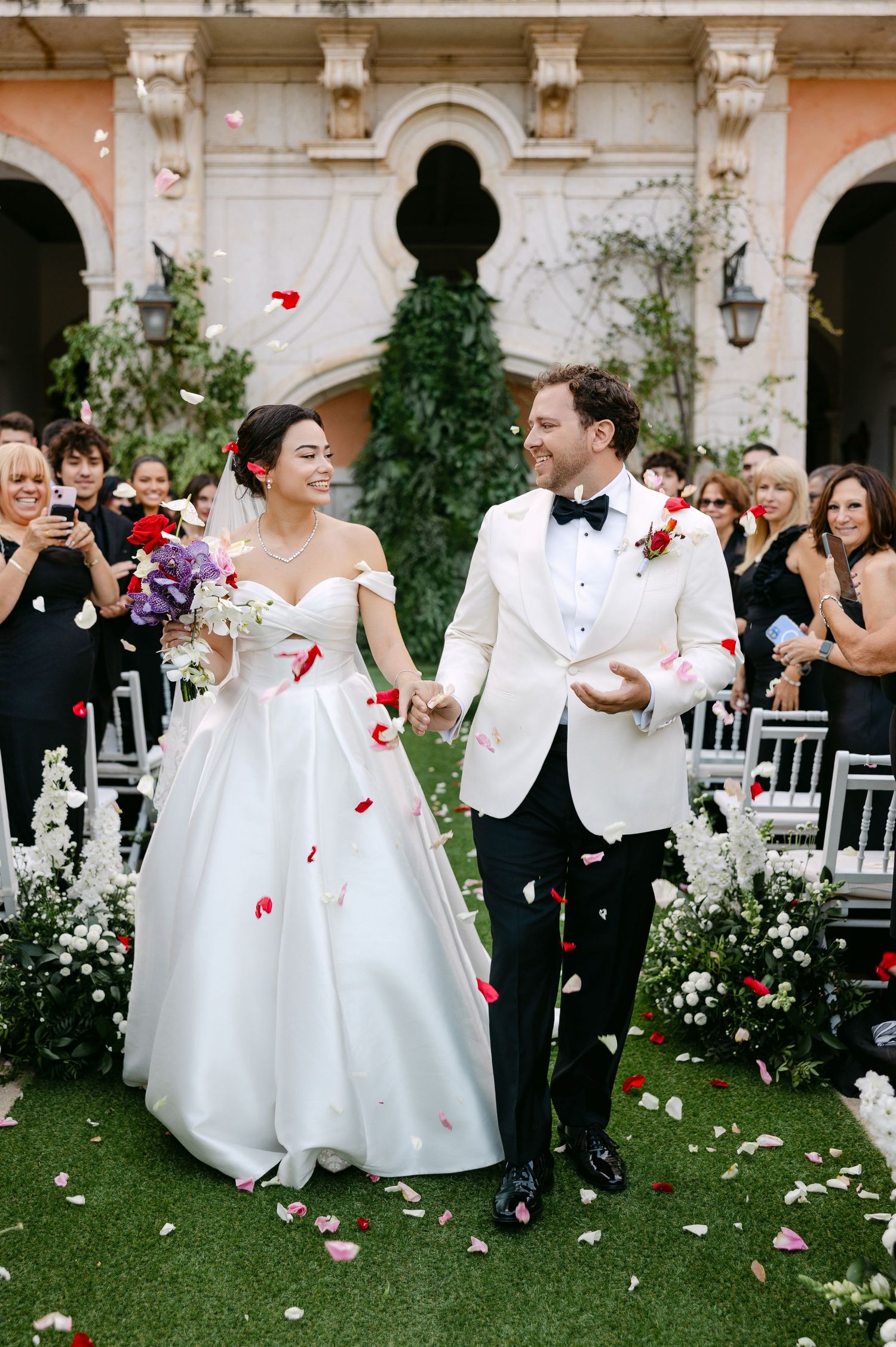 Wedding couple walking down aisle while guests throw rose petals in elegant garden ceremony setting.