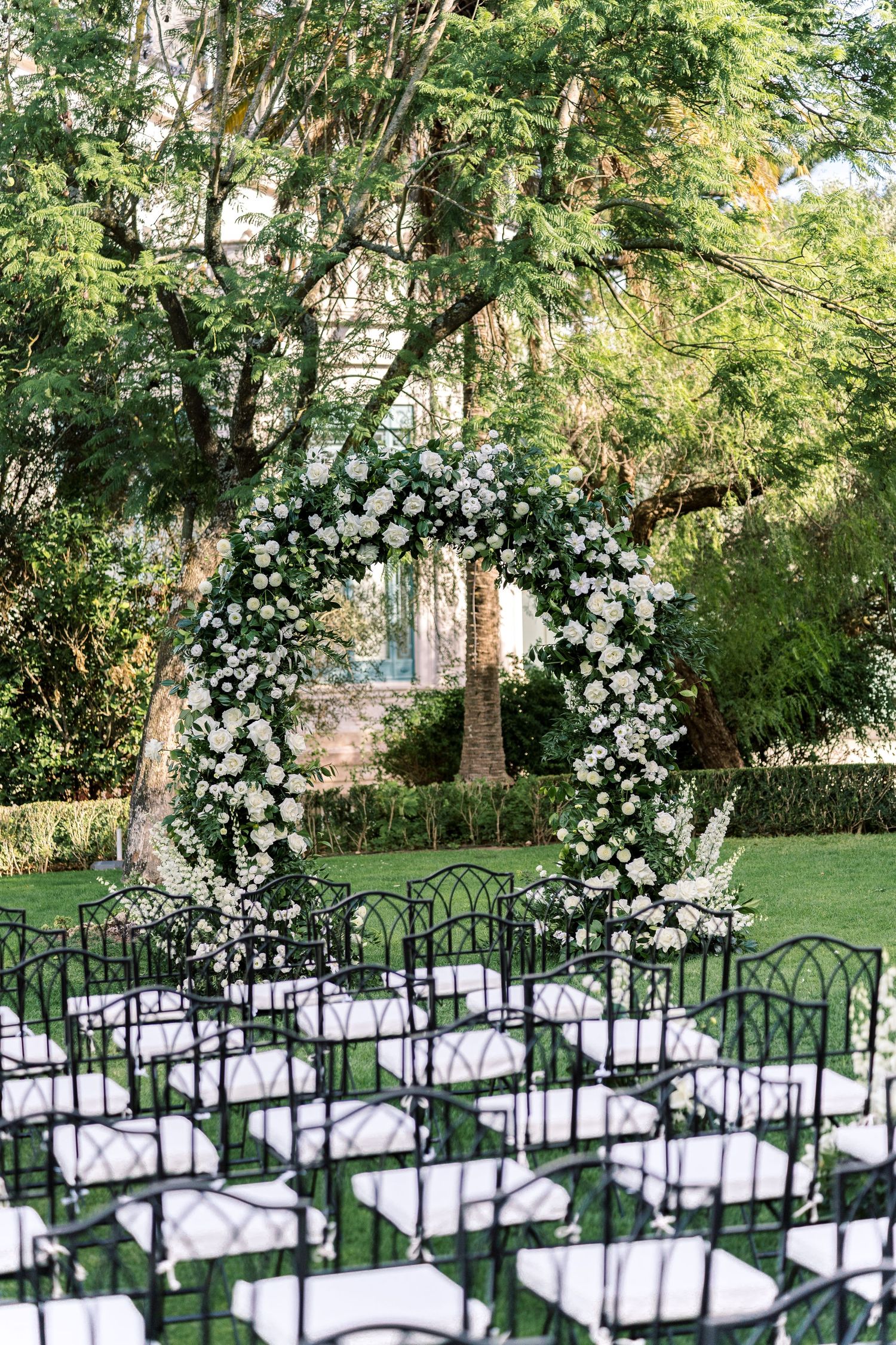 White floral wedding arch with rows of black chairs set up for outdoor garden ceremony.
