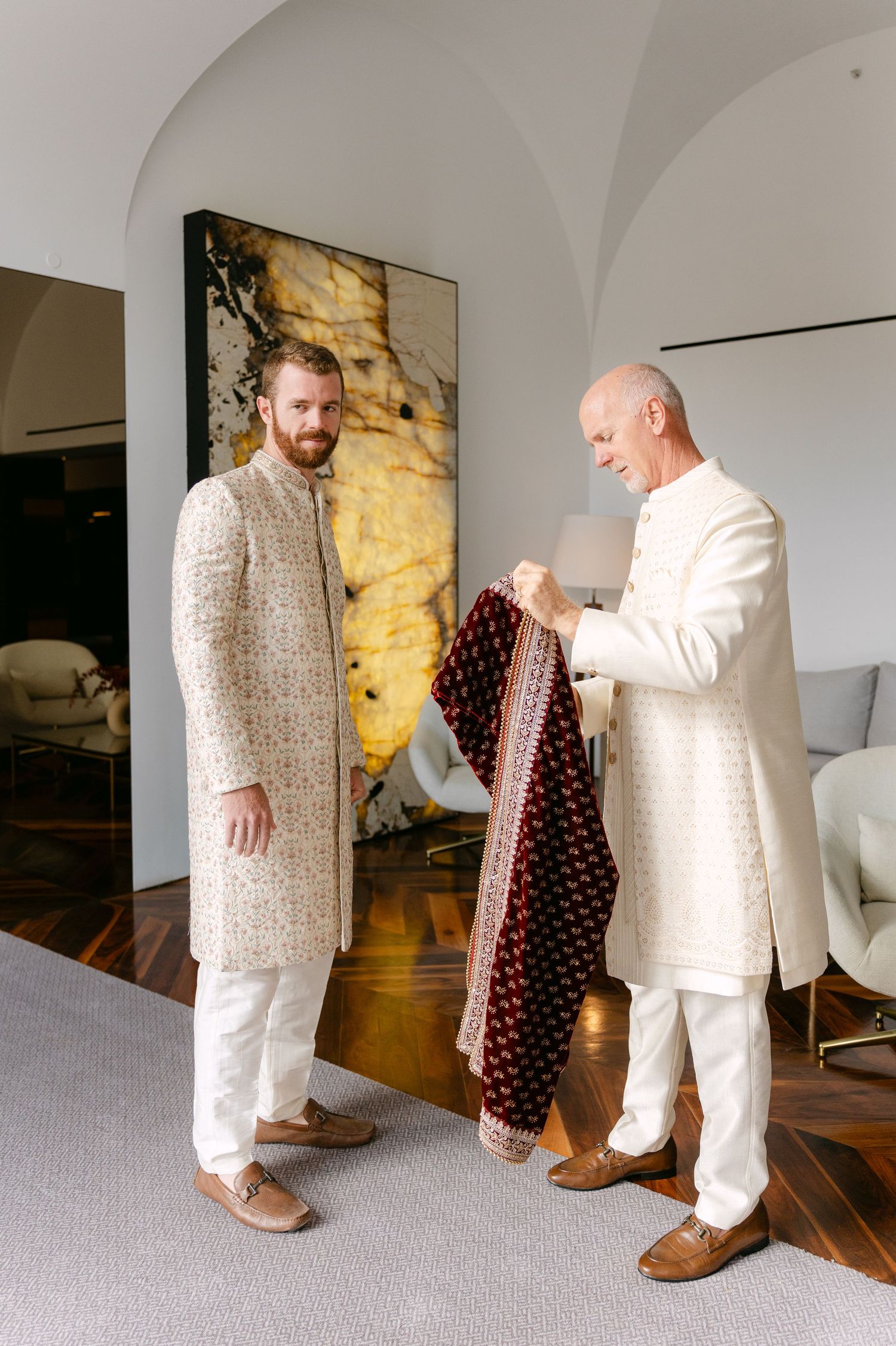 Two people in white and cream traditional Indian wedding attire getting ready indoors.