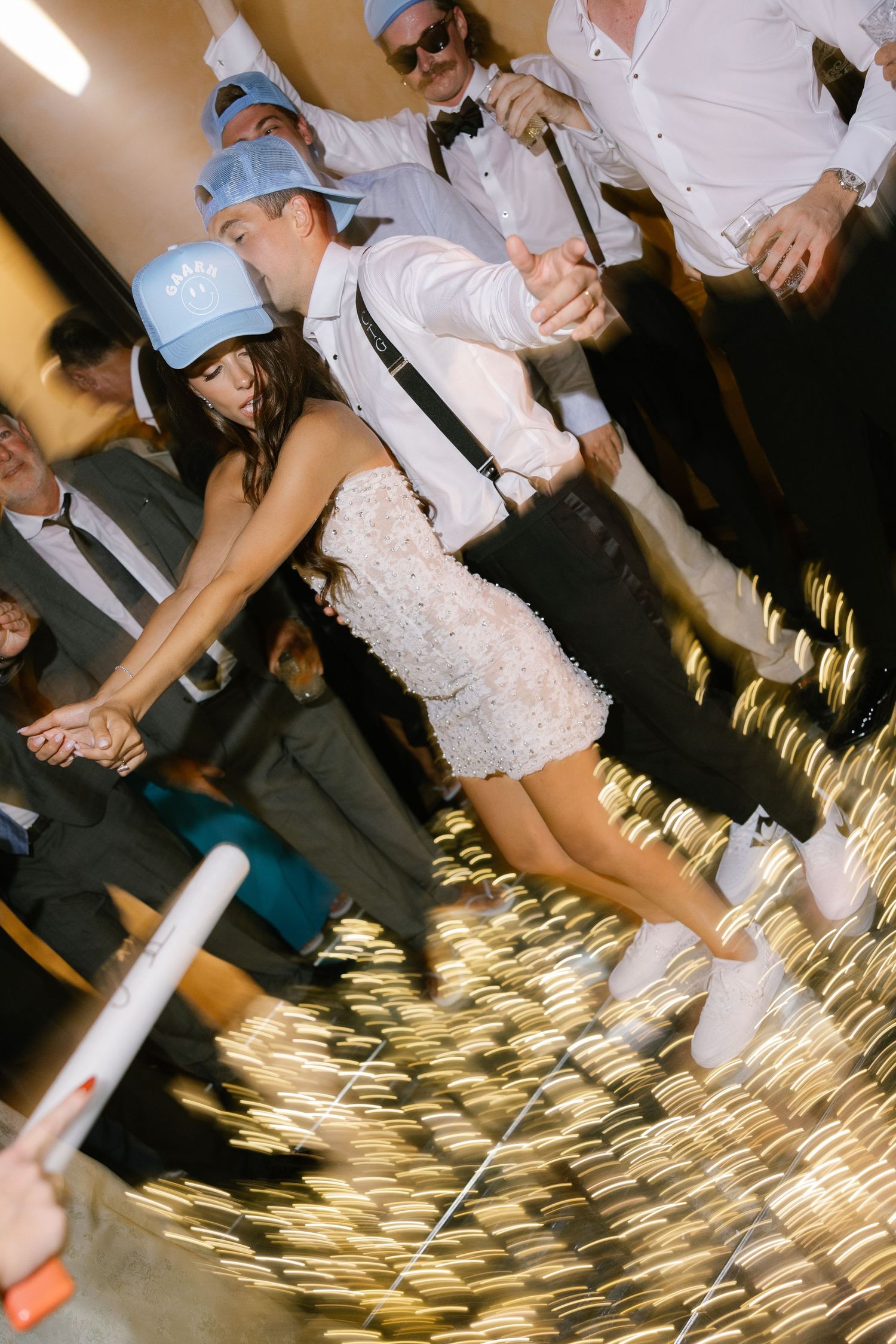 Wedding party dancing with sparkler effects and light blue baseball caps.