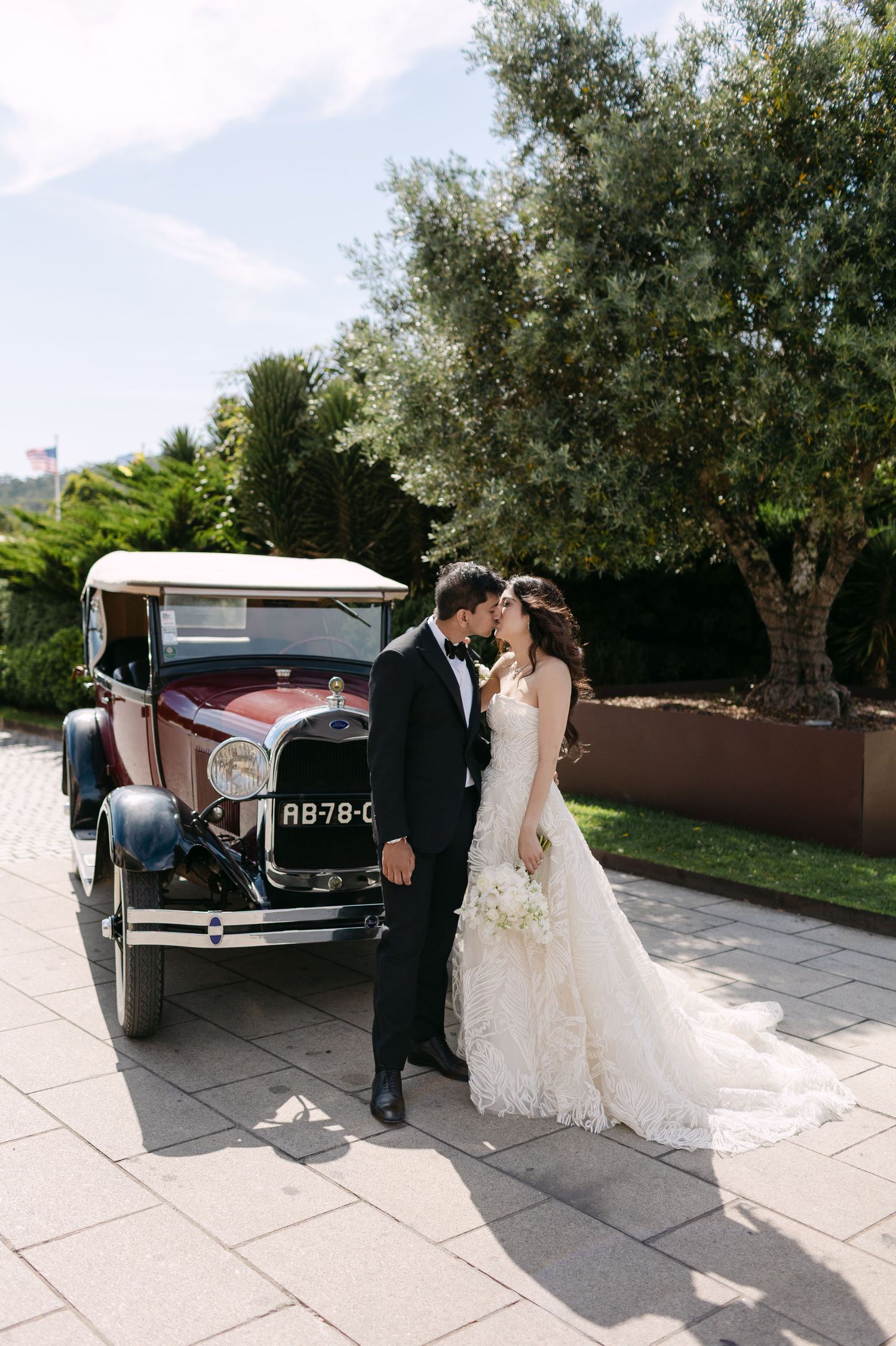 Elegant wedding couple shares intimate moments beside a vintage automobile in outdoor setting.