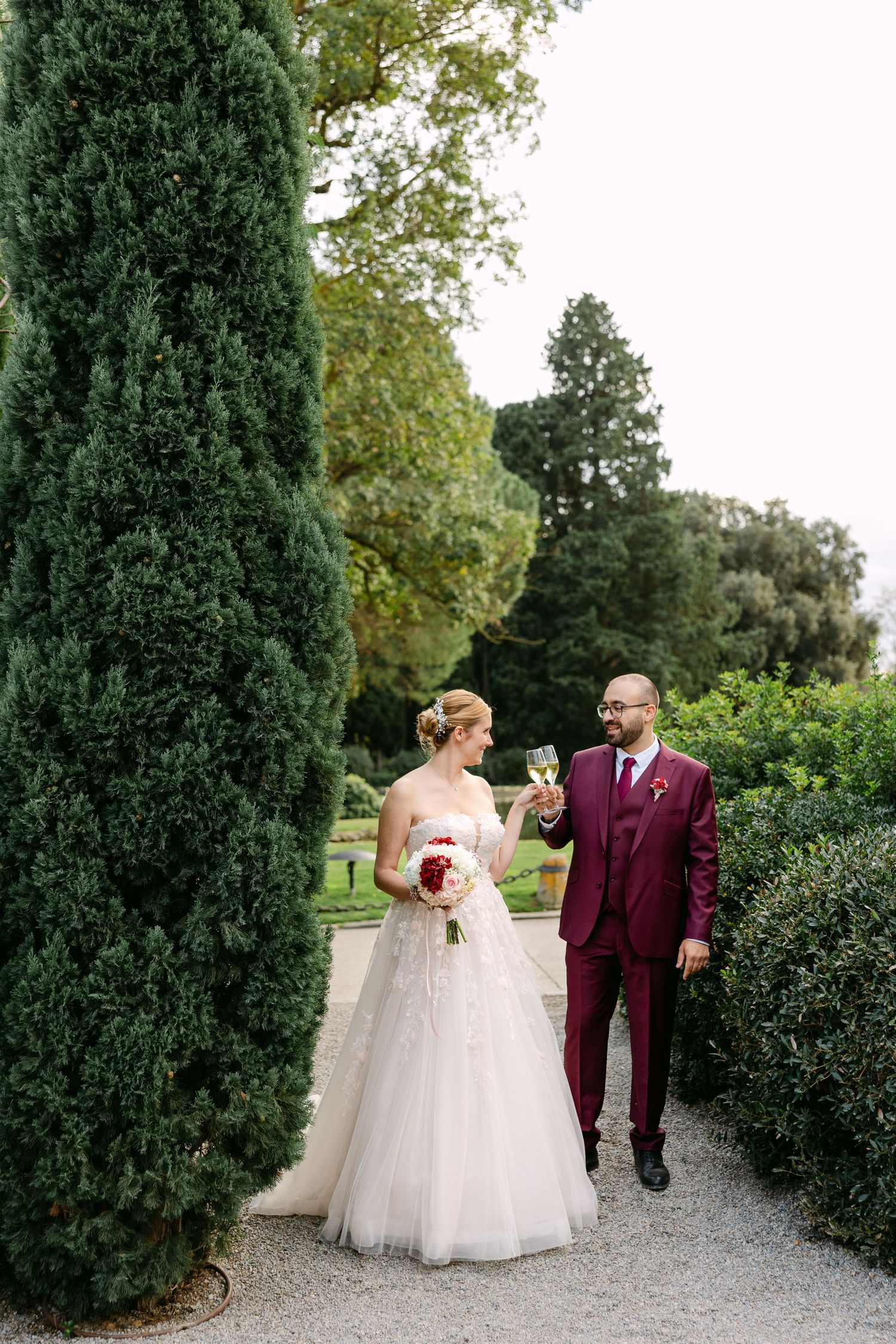 Couple walks along a garden path lined with tall cypress trees on their wedding day.