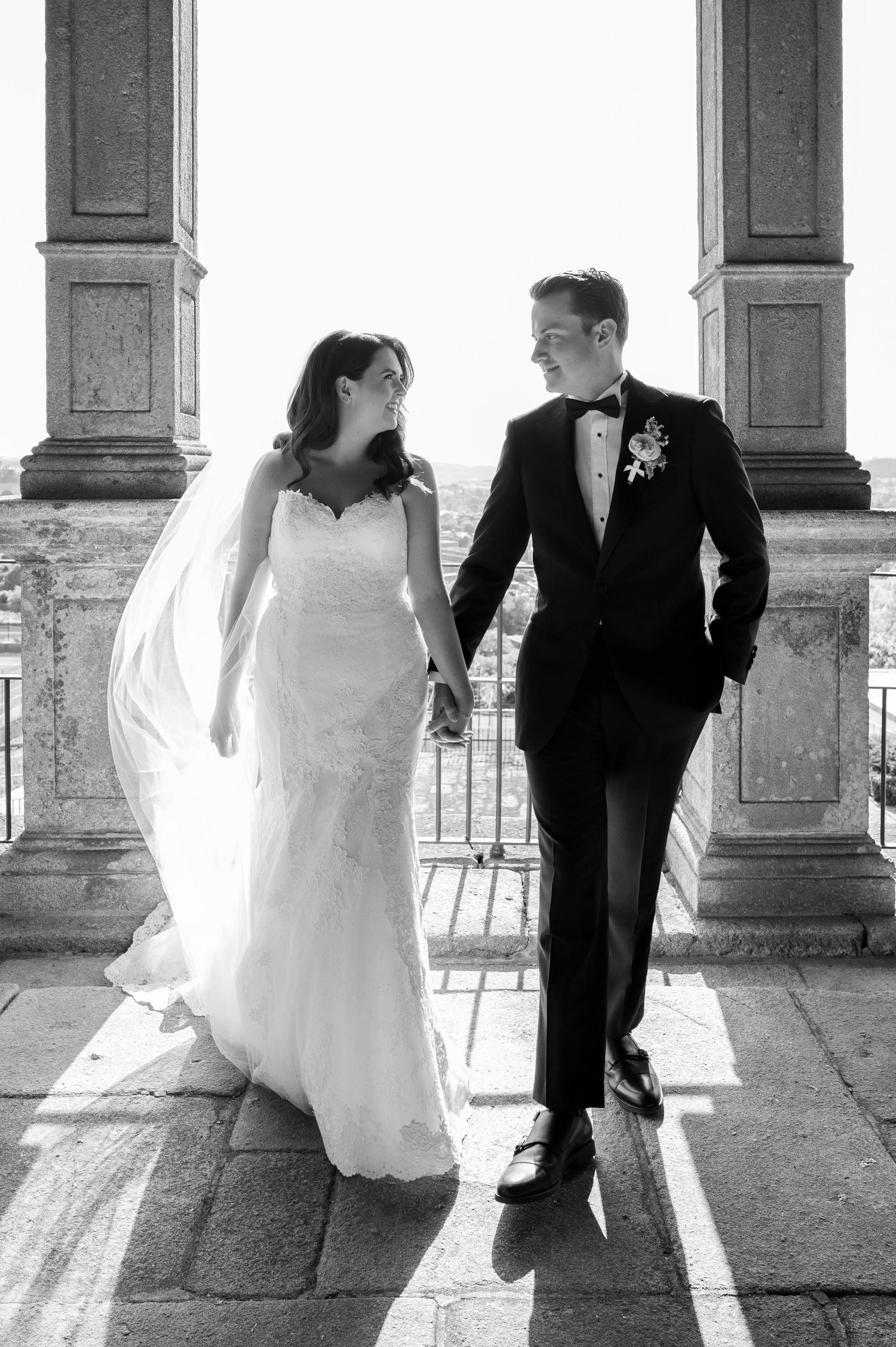Black and white portrait of couple in wedding attire by stone pillars.