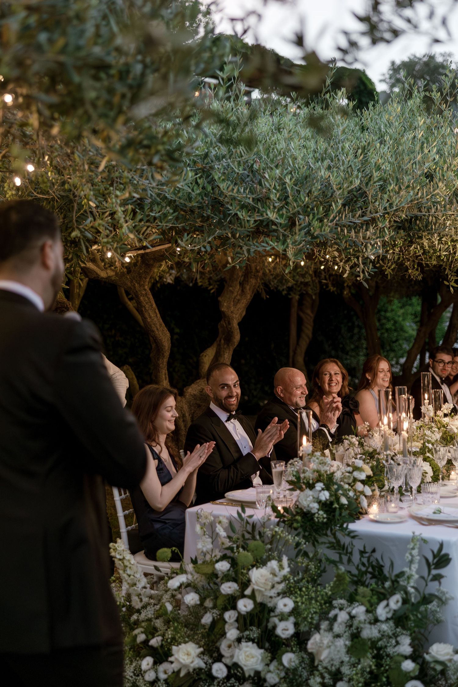 Guests applaud at an elegant outdoor wedding reception dinner under olive trees with white floral arrangements and string lights.