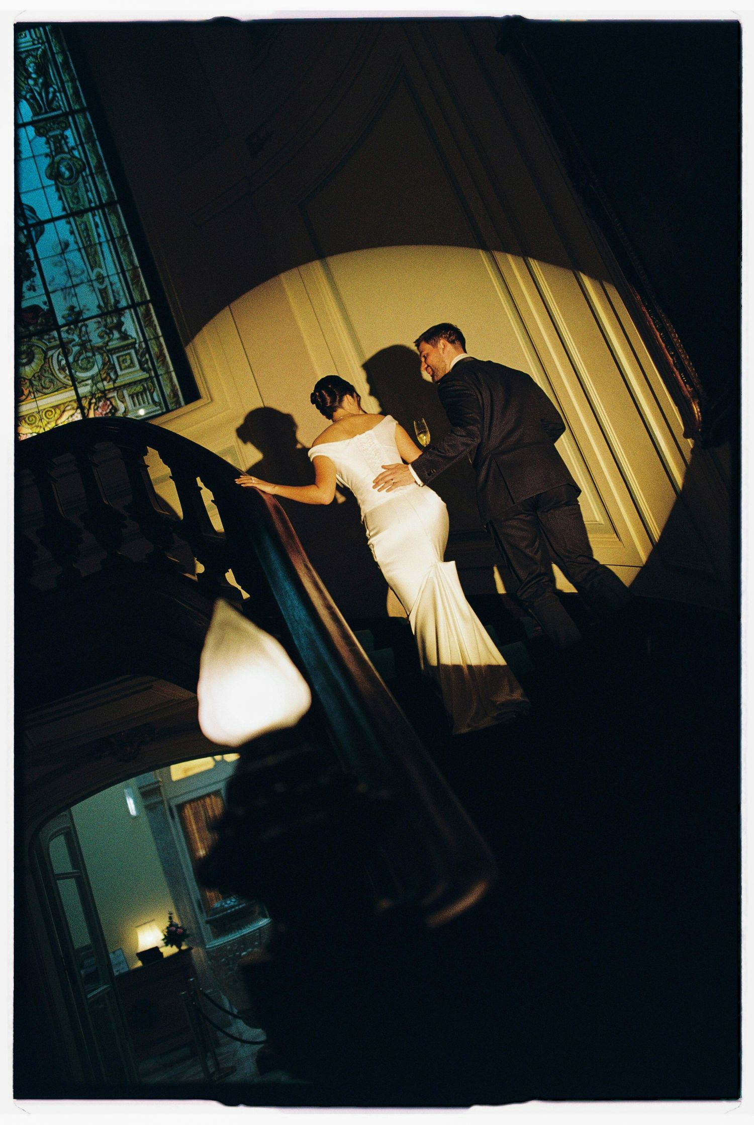 Bride in white mermaid gown and groom in black suit stand on elegant staircase with stained glass window.