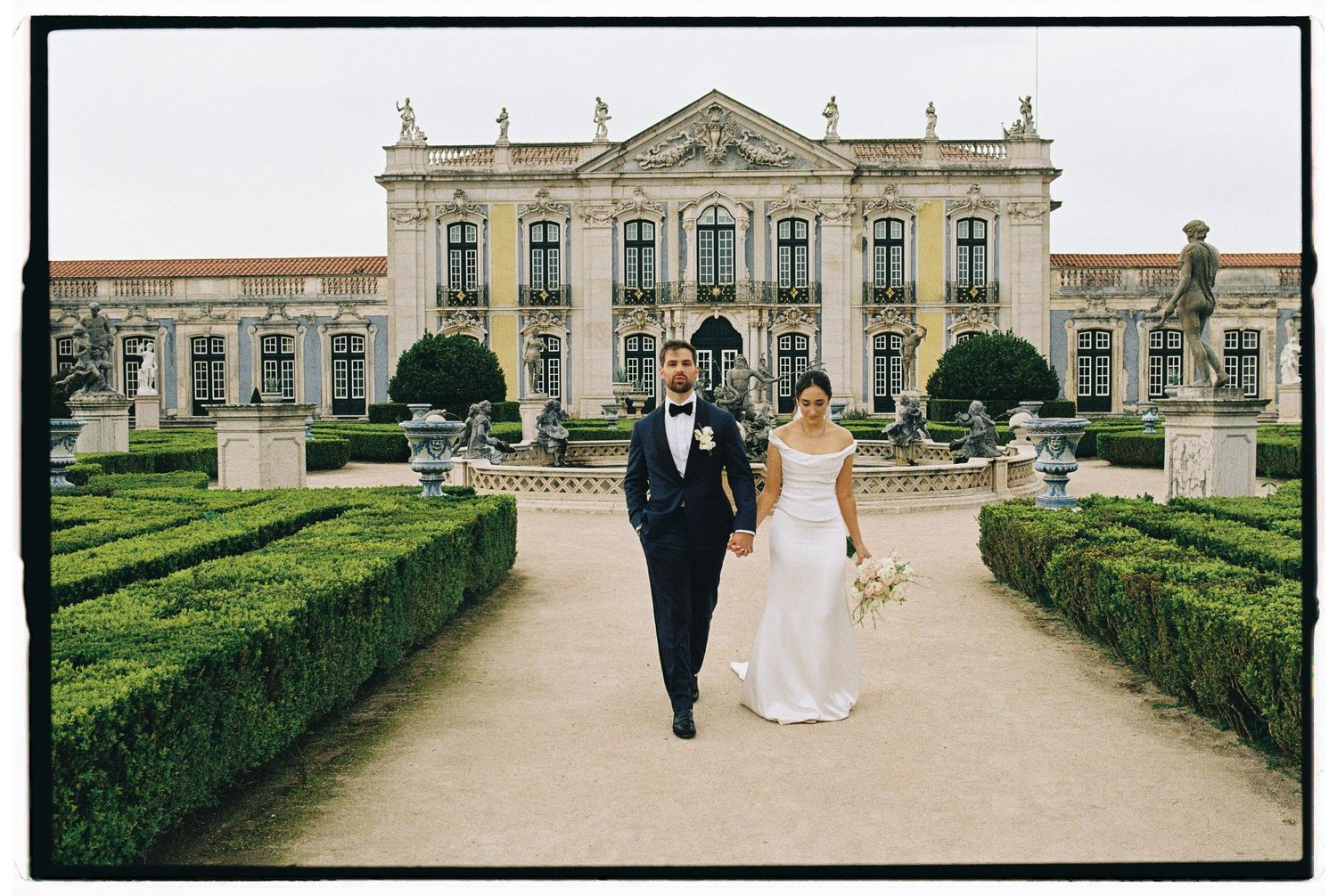Bride and groom walk through formal garden path before grand baroque palace on their wedding day.